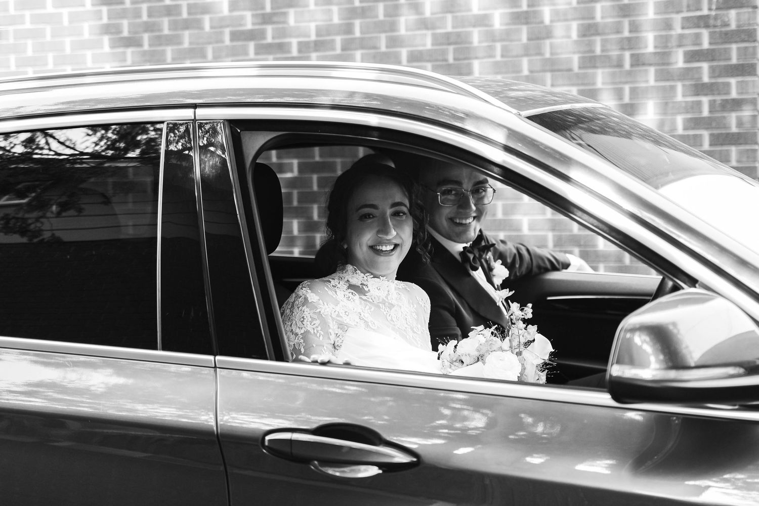 Black and white photo of a smiling bride holding flowers in a car with groom on wedding day.