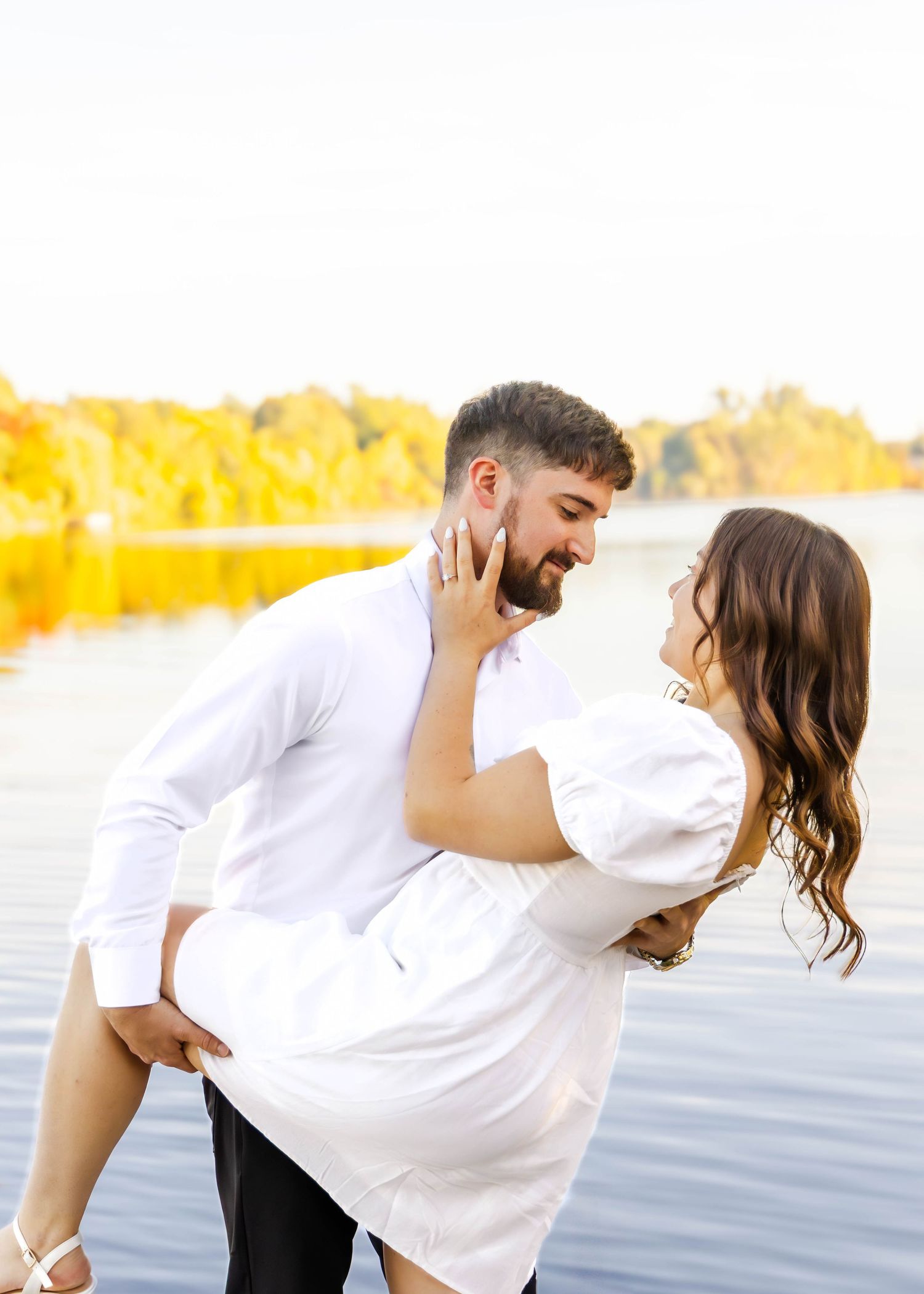Playful couple in white clothing embrace near a lake with golden fall foliage reflecting in the water.