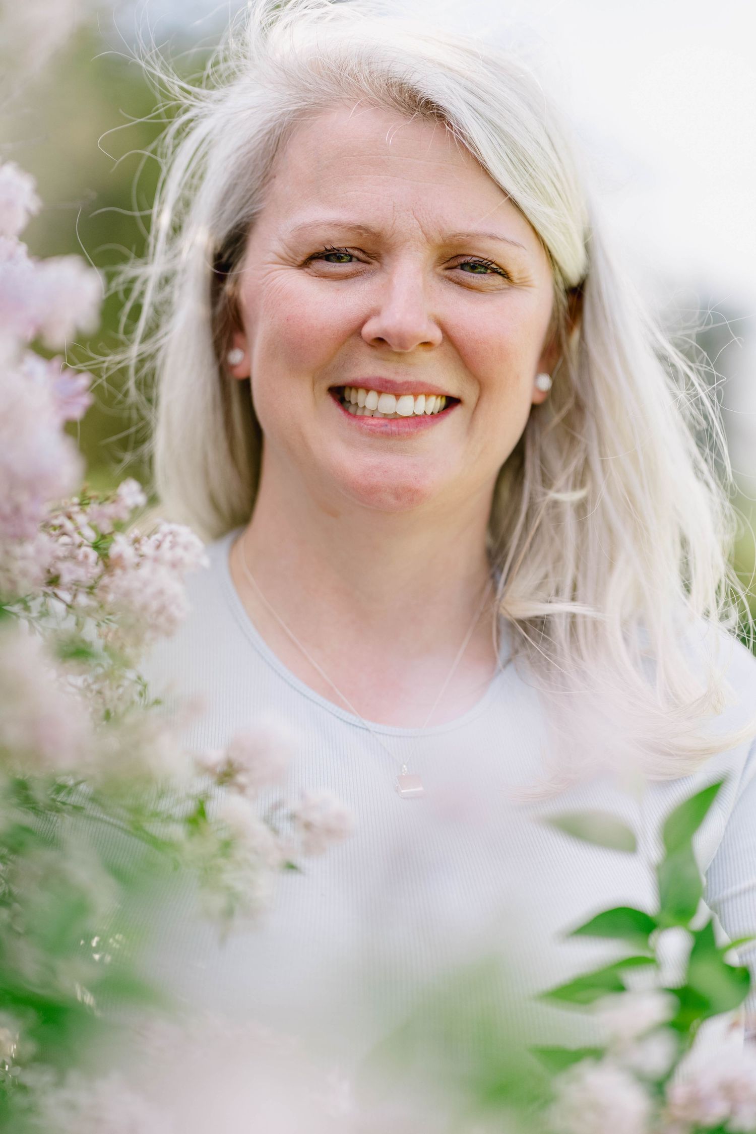 Smiling blonde woman surrounded by soft pink blooms in a bright outdoor garden setting.