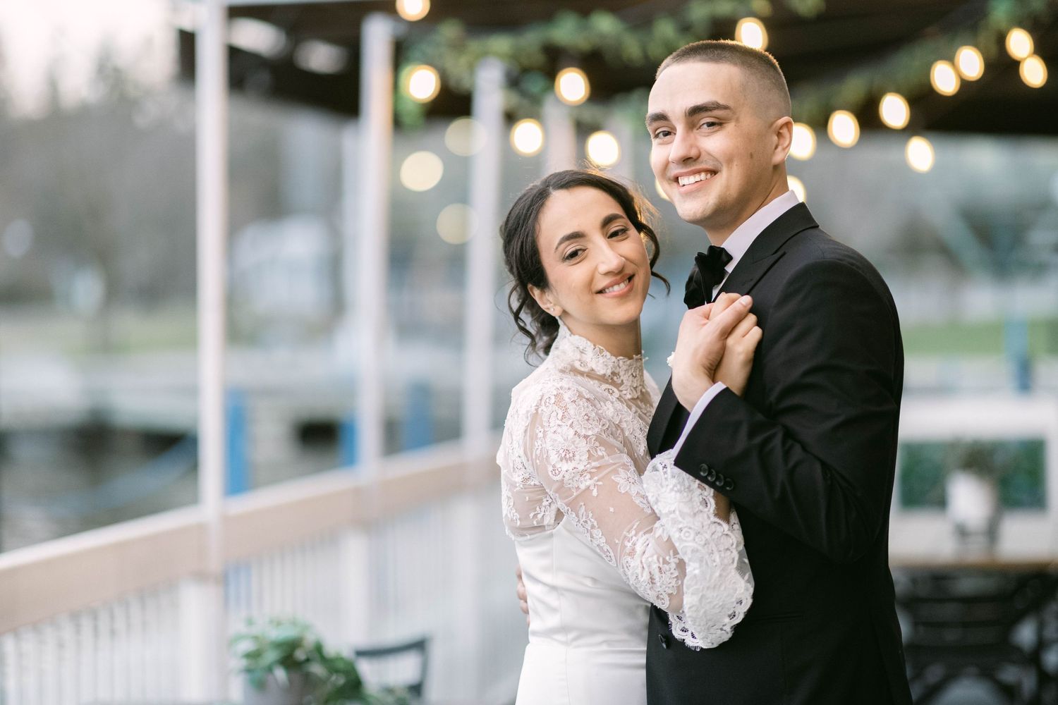 A bride and groom share a romantic first dance outdoors at a waterfront wedding venue with warm string lights.