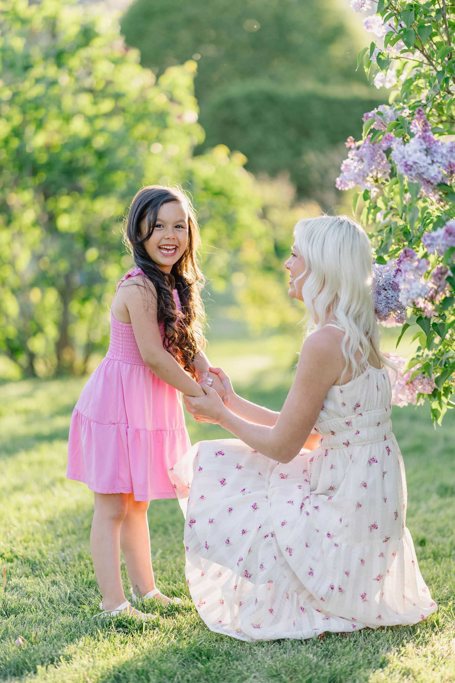 A blonde woman kneels holding hands with a smiling girl in pink dress near lilac blooms in sunny garden.