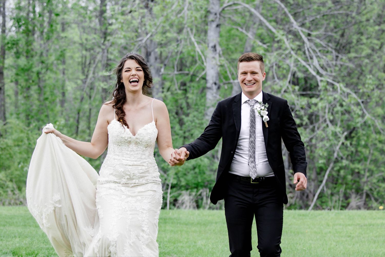 Newlyweds joyfully run through a lush green forest setting in formal wedding attire.