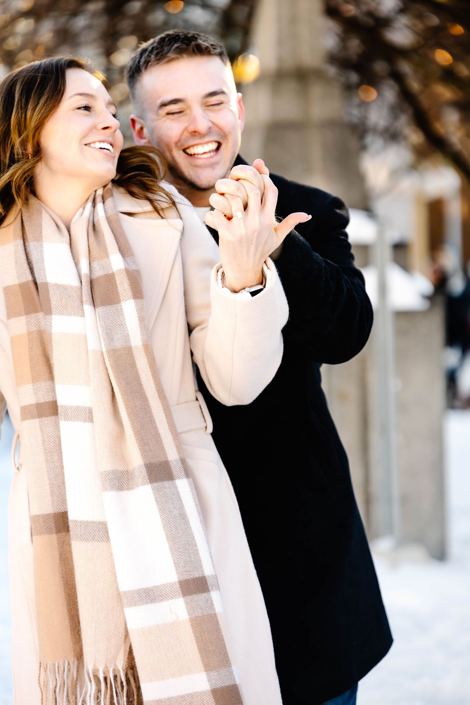 A couple shares joyful laughter while standing together in a snowy winter setting wearing a plaid scarf and black coat.