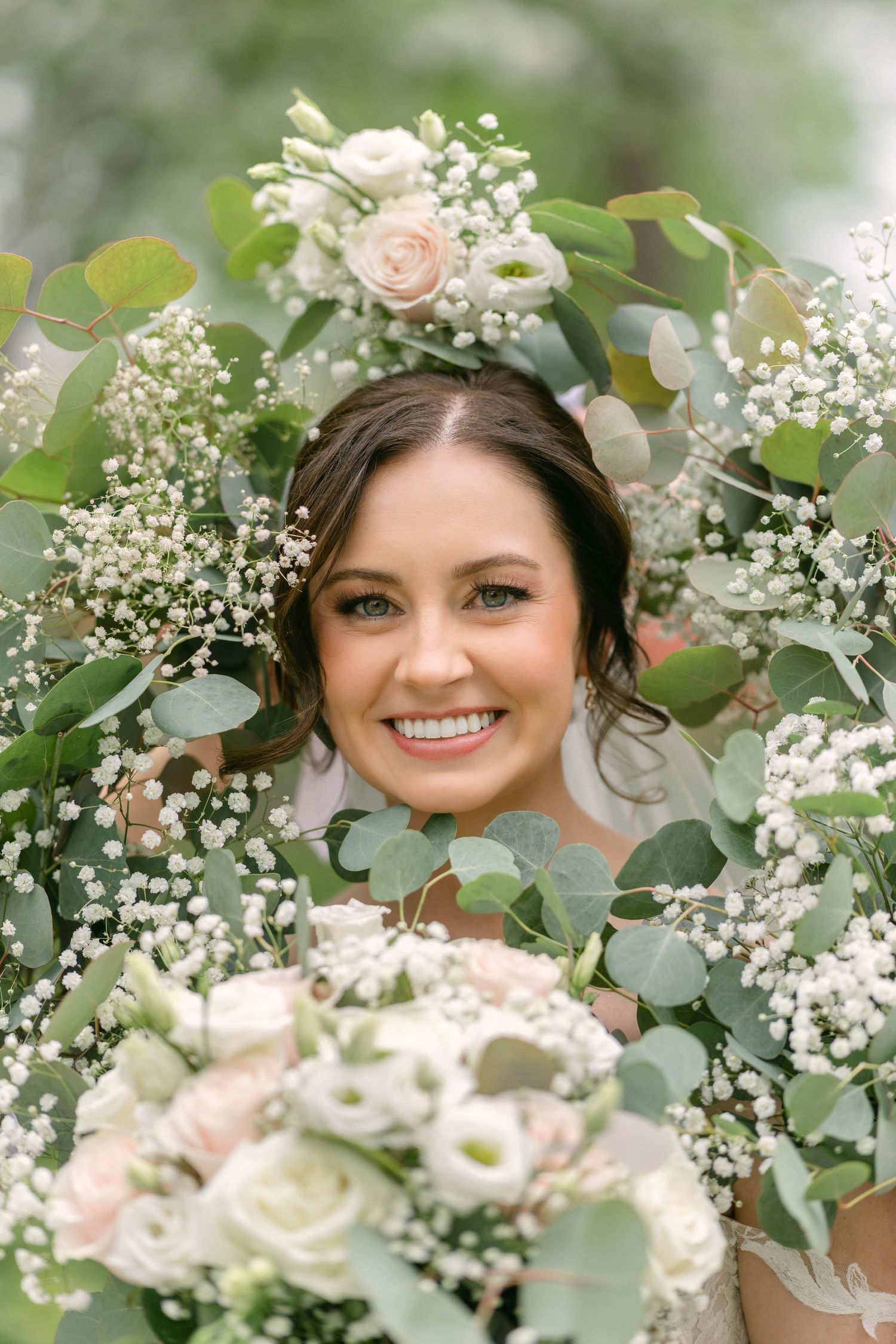 Smiling bride surrounded by lush eucalyptus, baby's breath, and blush roses bouquet on her wedding day.