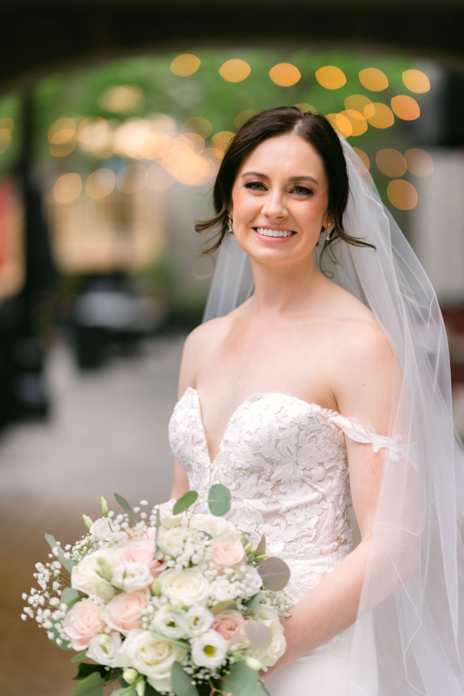 Smiling bride in lace off-shoulder wedding gown holds blush and white bouquet with bokeh lights behind her.