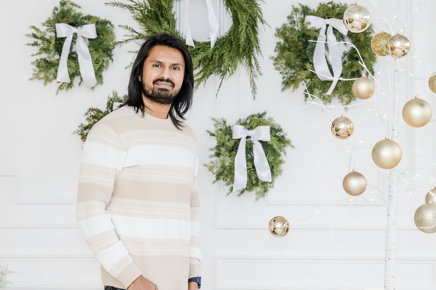 Minimal holiday backdrop with evergreen wreaths and gold ornaments against white wall.