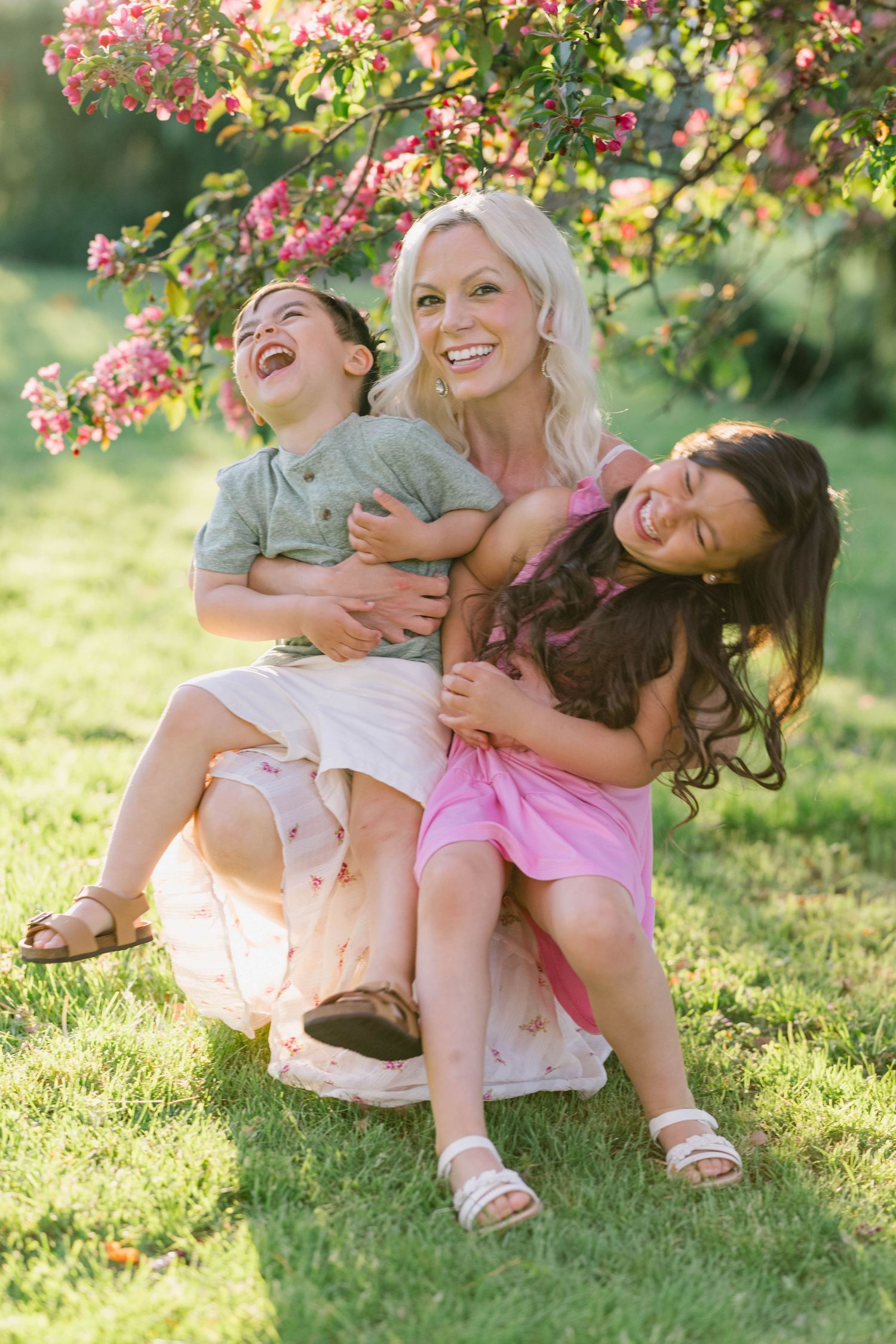 A joyful blonde mother sits on grass with two laughing children on her lap under pink flowering tree.