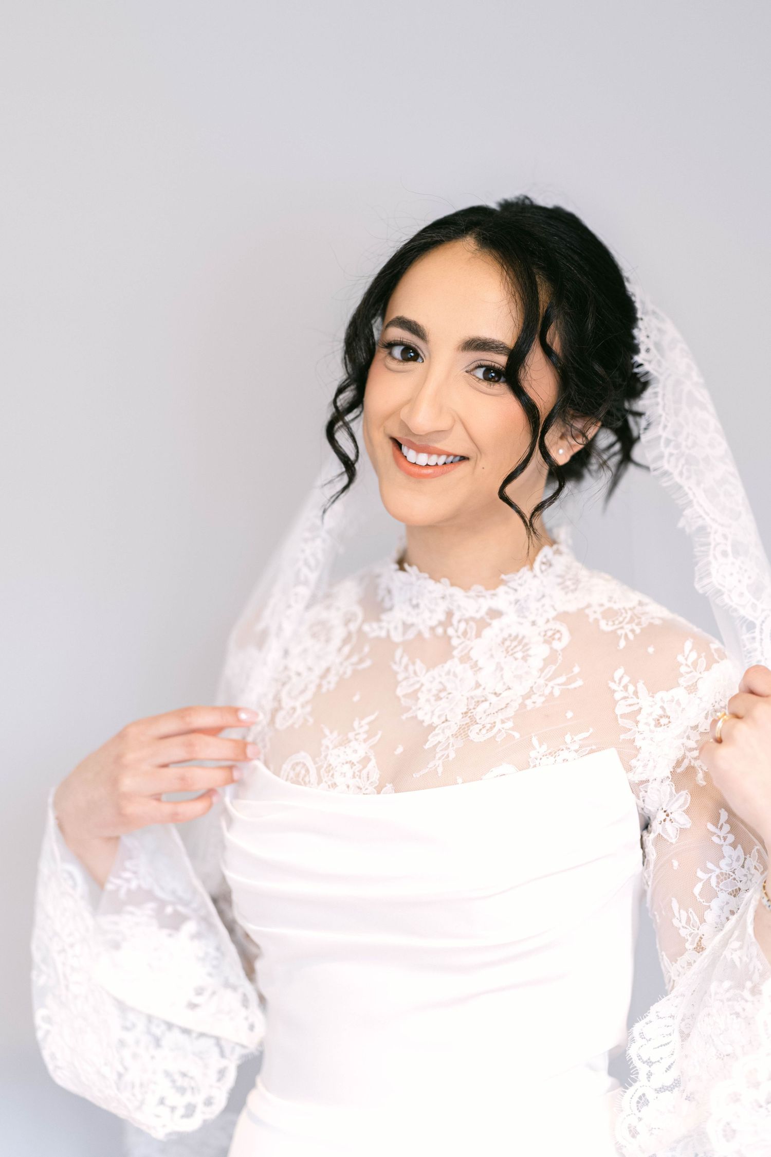 Smiling bride in white lace wedding dress and veil with dark curly updo hair against gray background.