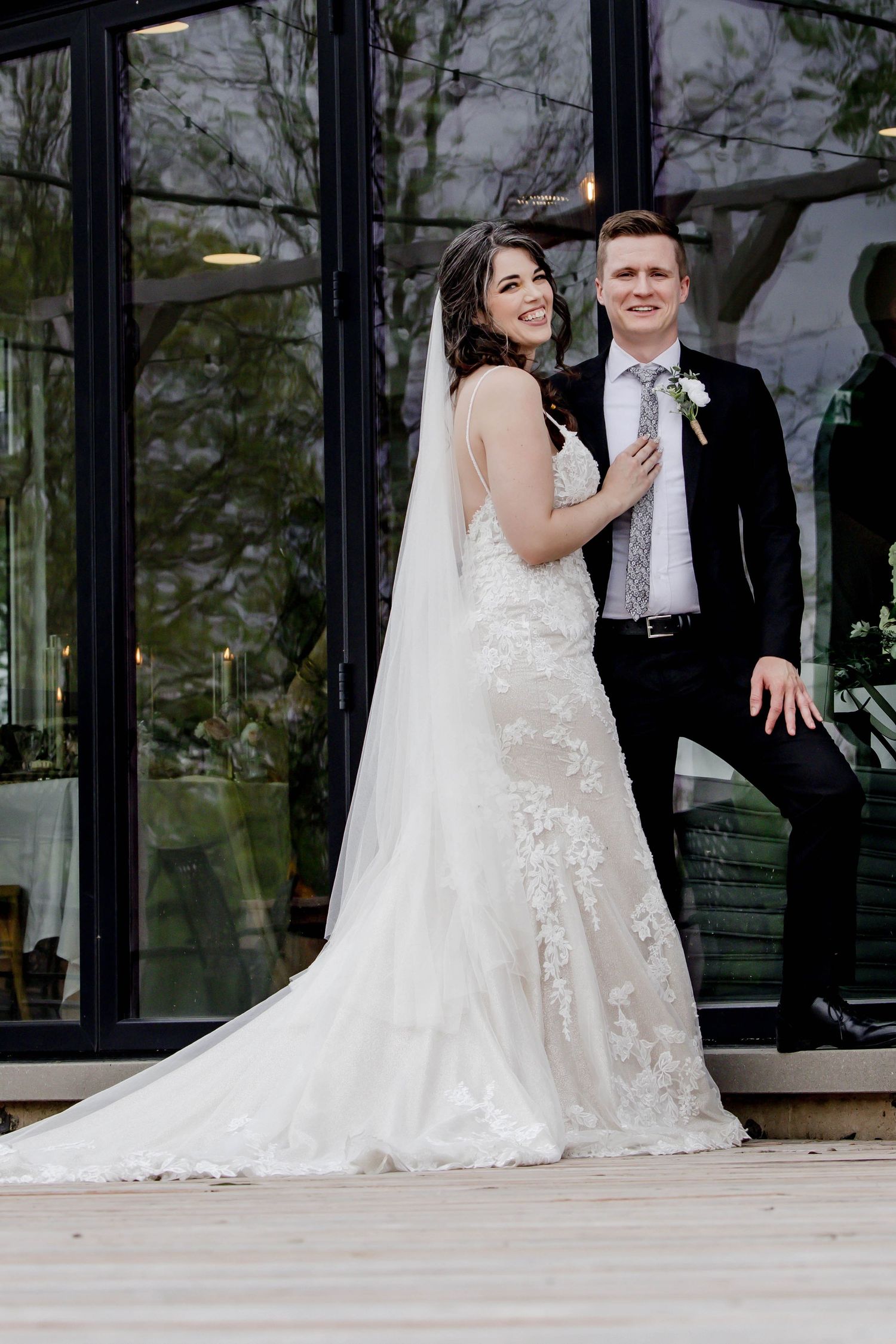 A couple shares a playful moment during their wedding photoshoot on a wooden deck with glass windows in the background.