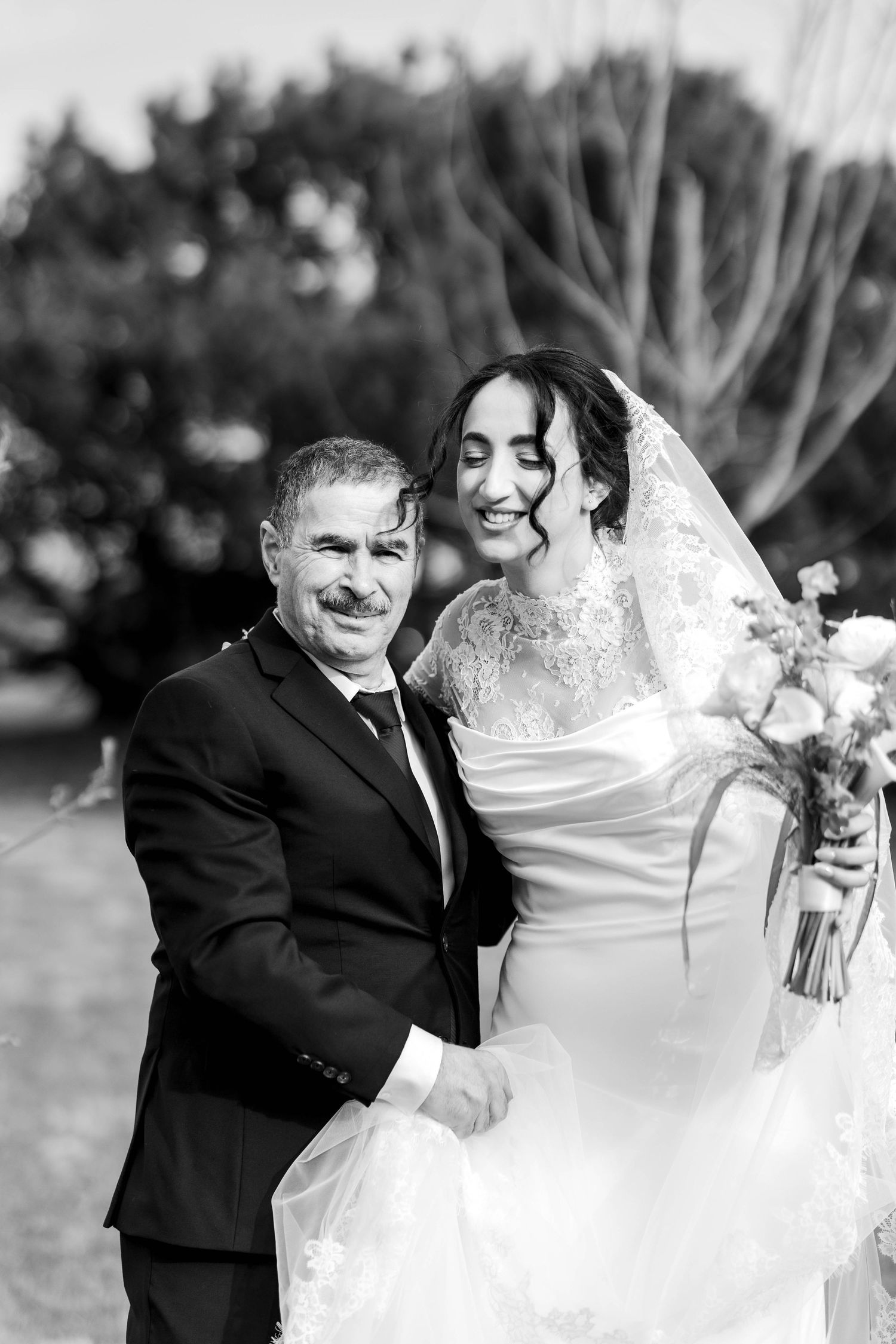 Black and white photo of a smiling bride in a lace veil holding bouquet beside a suited man at an outdoor wedding.