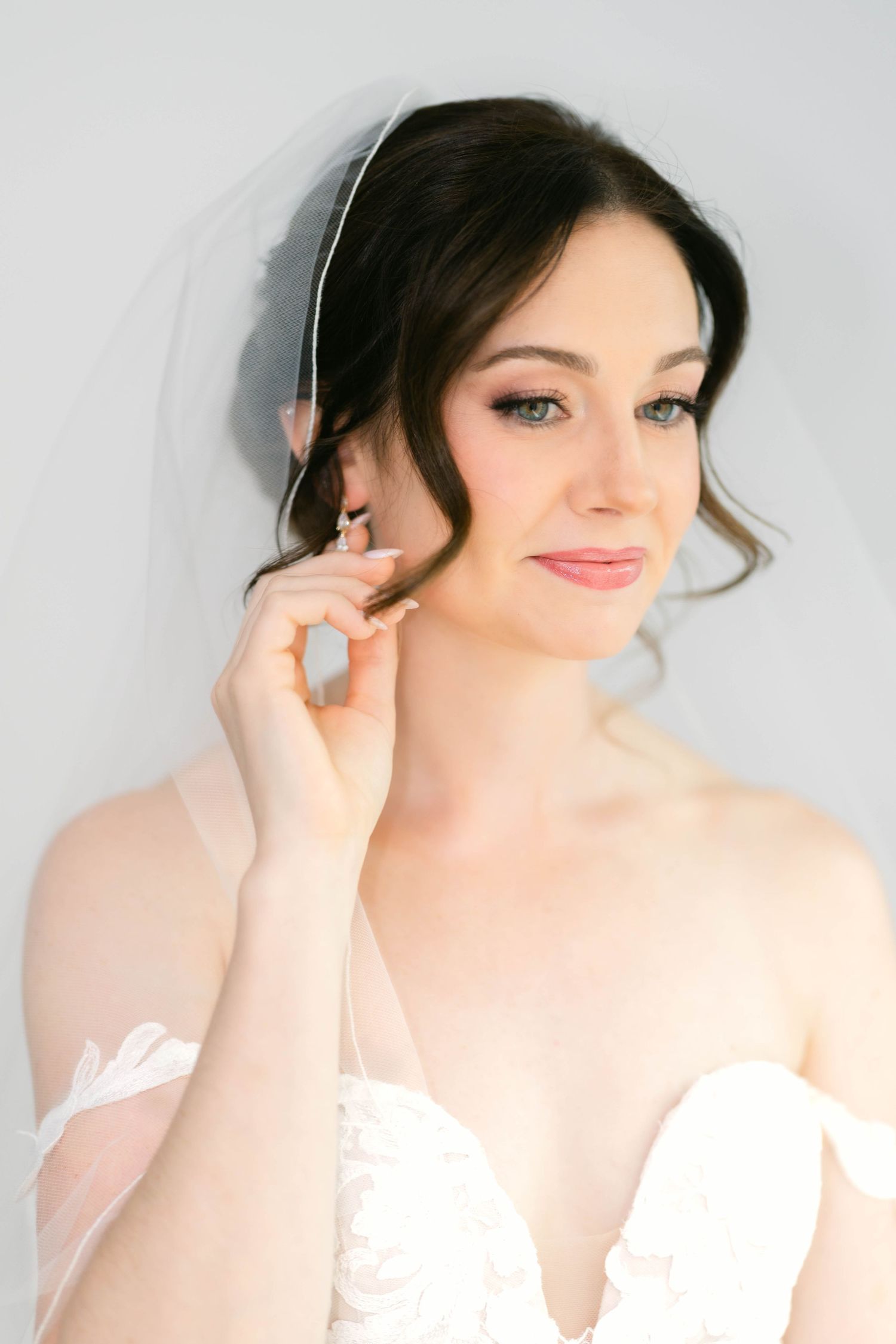 Elegant bride in white strapless gown and veil adjusting earring, with soft bridal makeup and dark updo hairstyle.