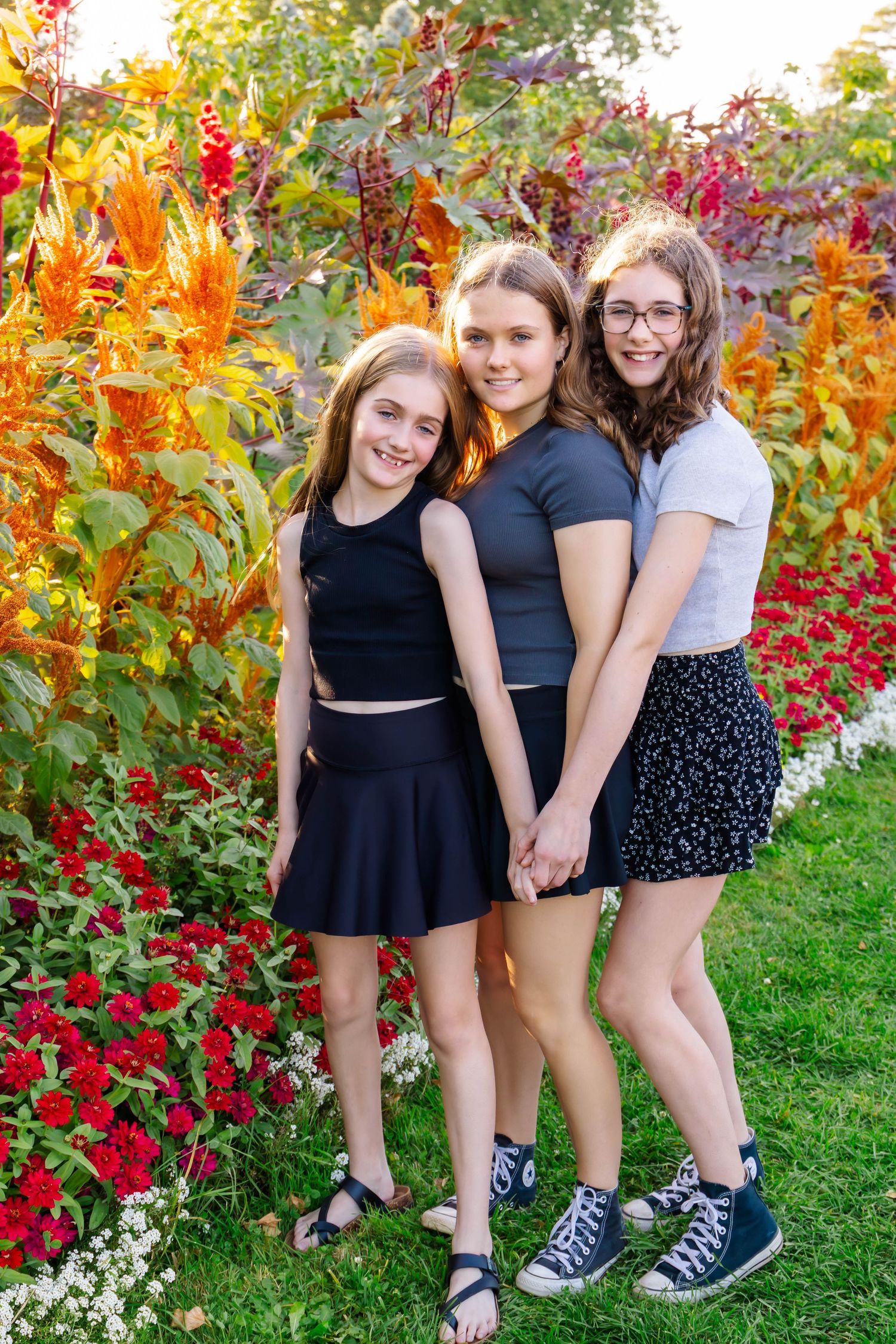 Friends stand together in coordinated black outfits against a backdrop of colorful autumn leaves and flowers.