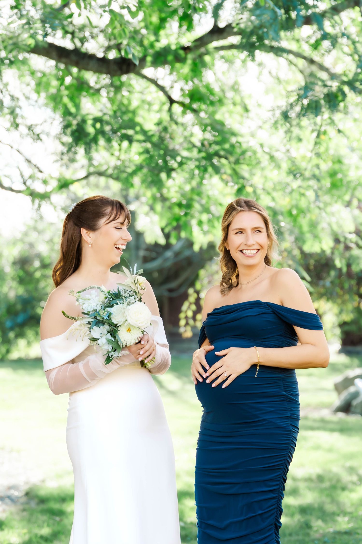A wedding party wearing white and navy blue dresses poses together in a garden setting under green trees.