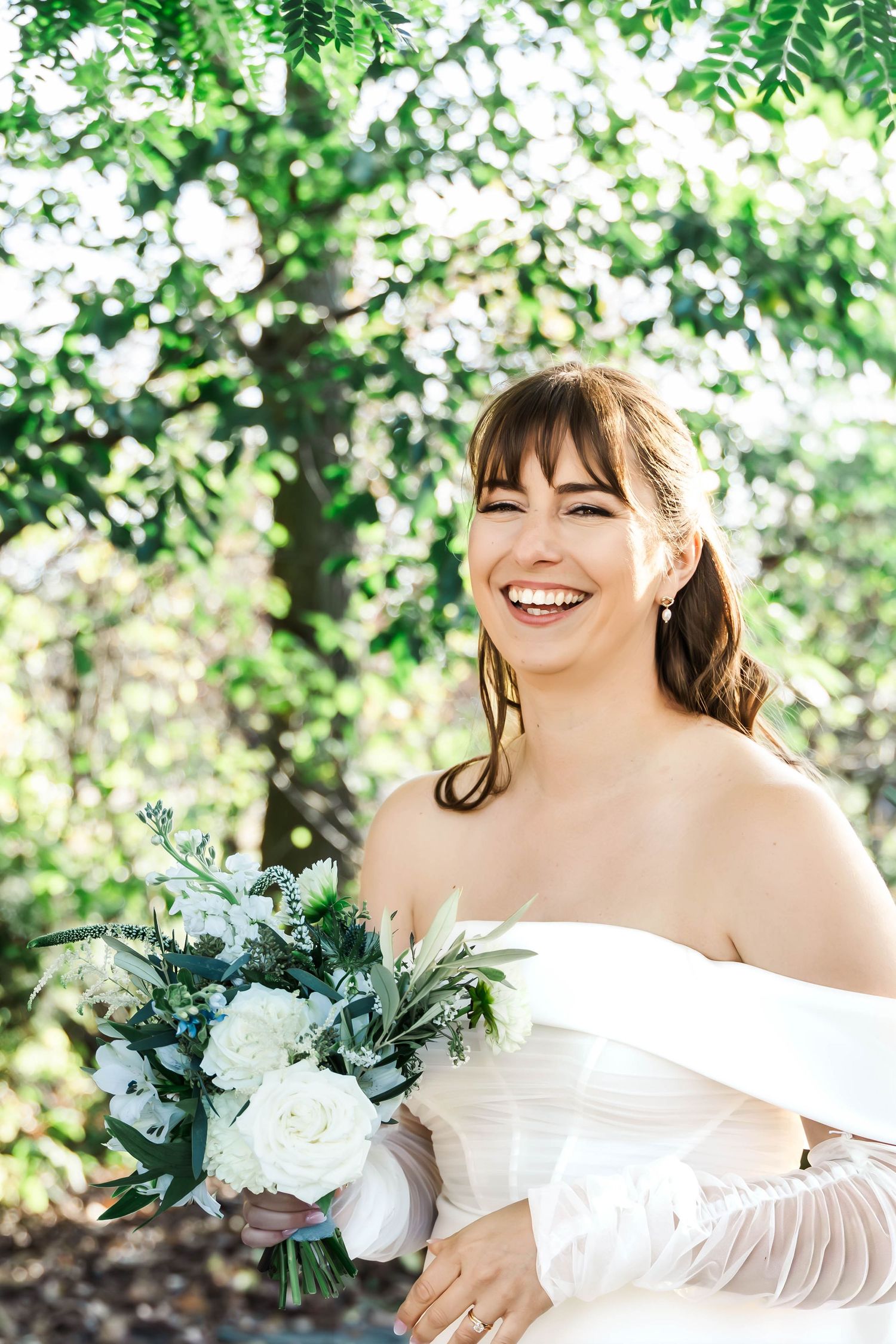 A bride in an off-shoulder white wedding dress smiles joyfully while holding a blue and white floral bouquet outdoors.