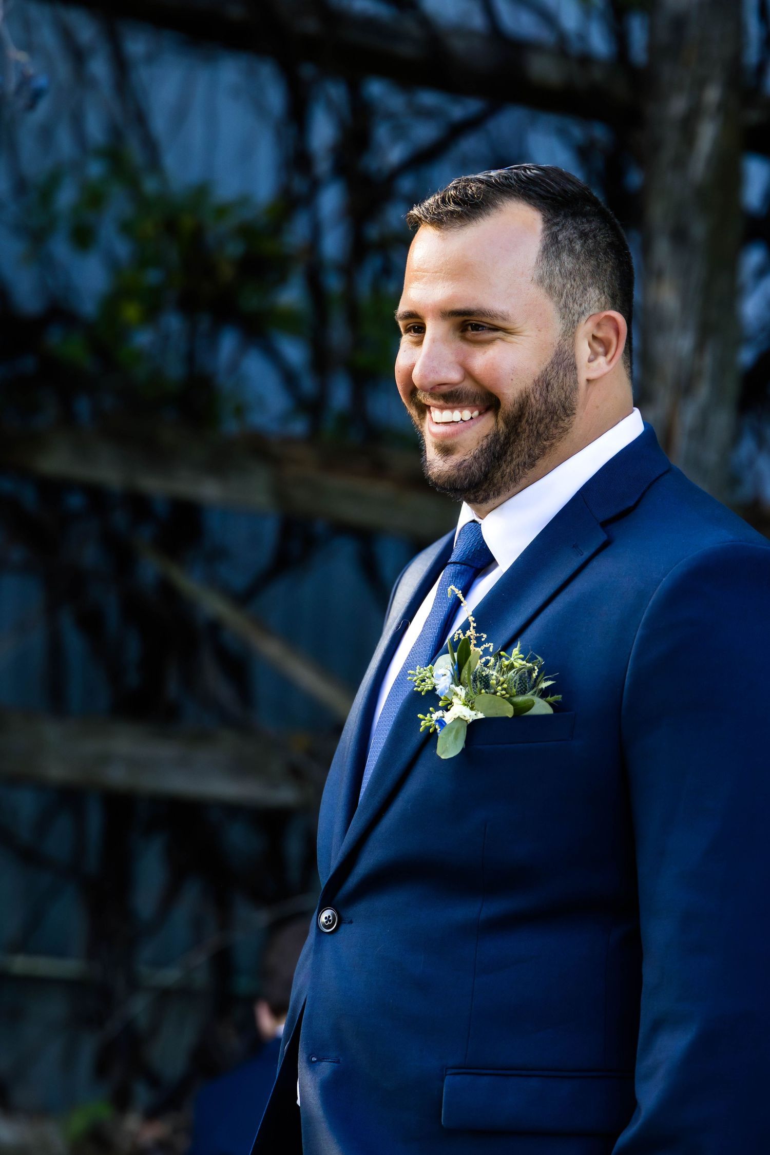 A groom in a classic navy suit with boutonniere smiles warmly during an outdoor wedding.