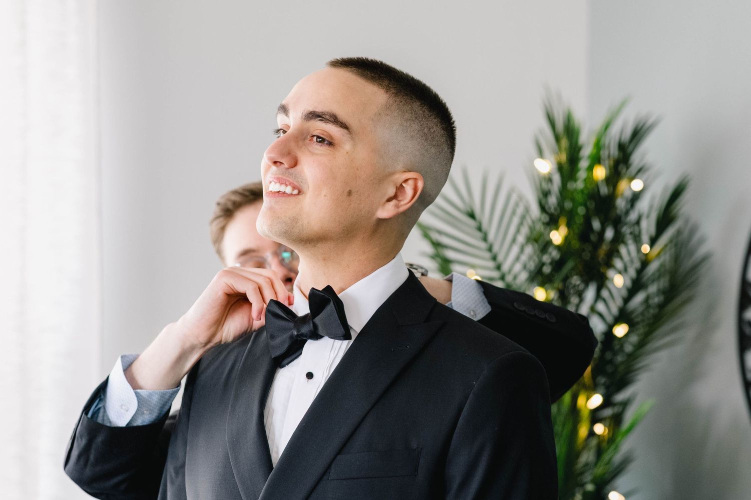 Young groom in black tuxedo and bow tie being helped by groomsman while smiling in bright room with tropical plant.