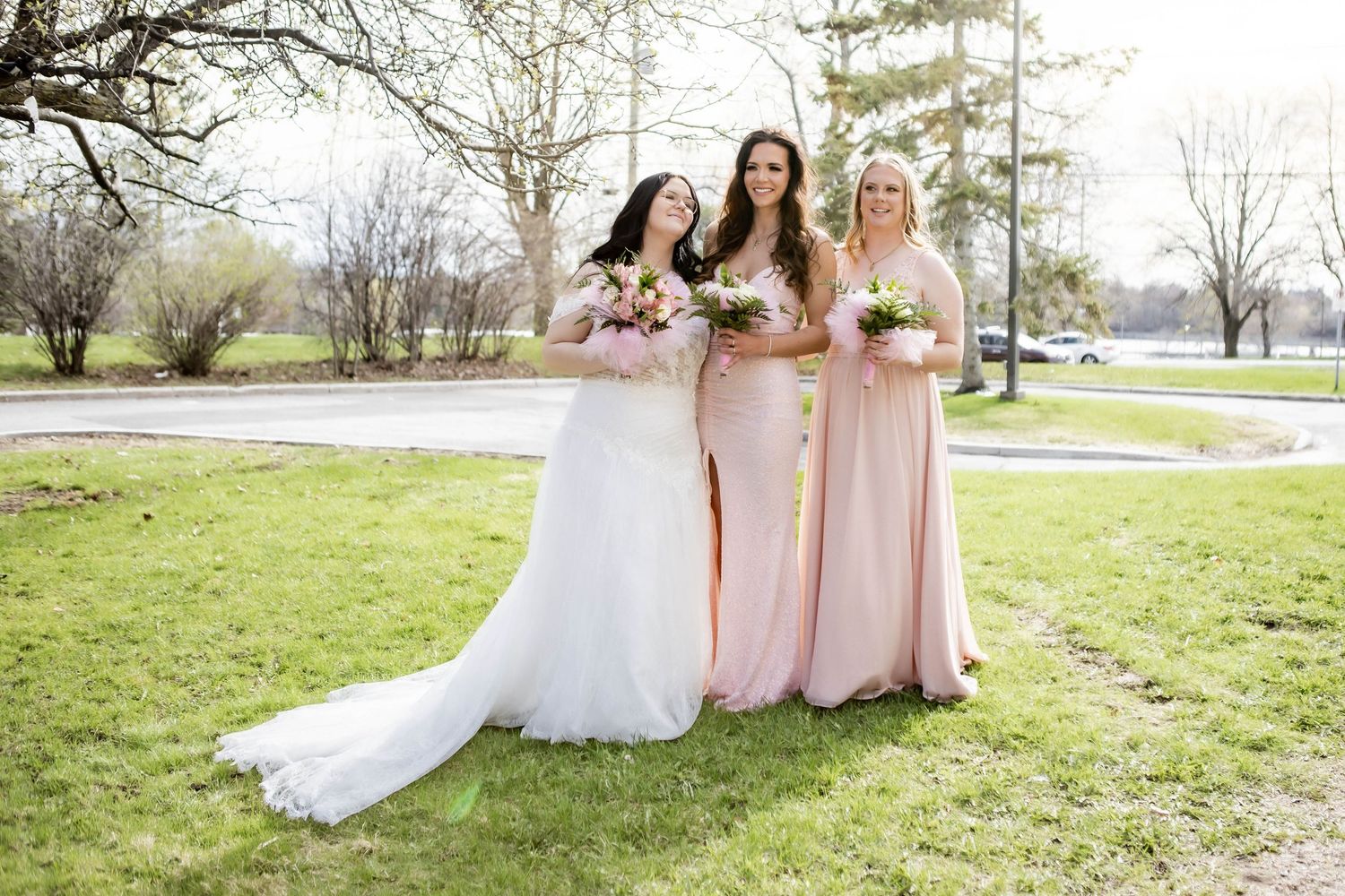 Bridal party in pink and white dresses poses on grass with spring trees in background.