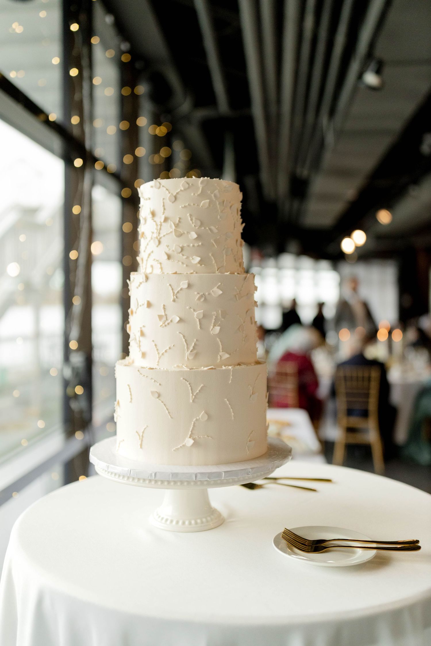 Elegant three-tier white wedding cake with floral details on white cake stand with gold utensils.
