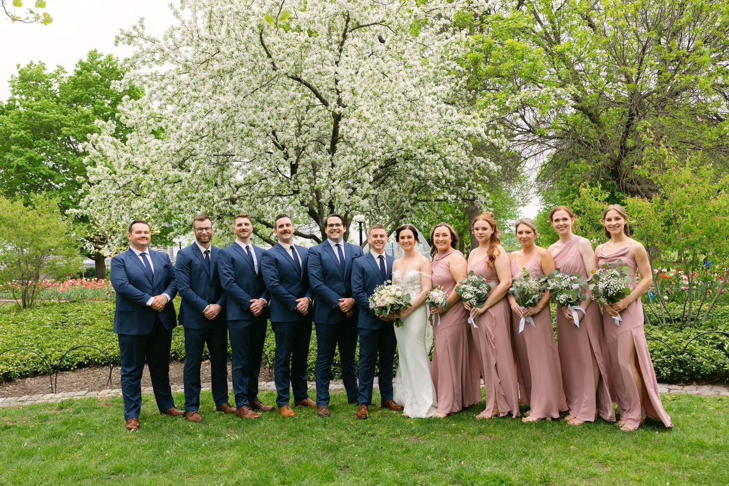 Wedding party in navy suits and blush gowns pose before blooming white trees in a lush garden setting.