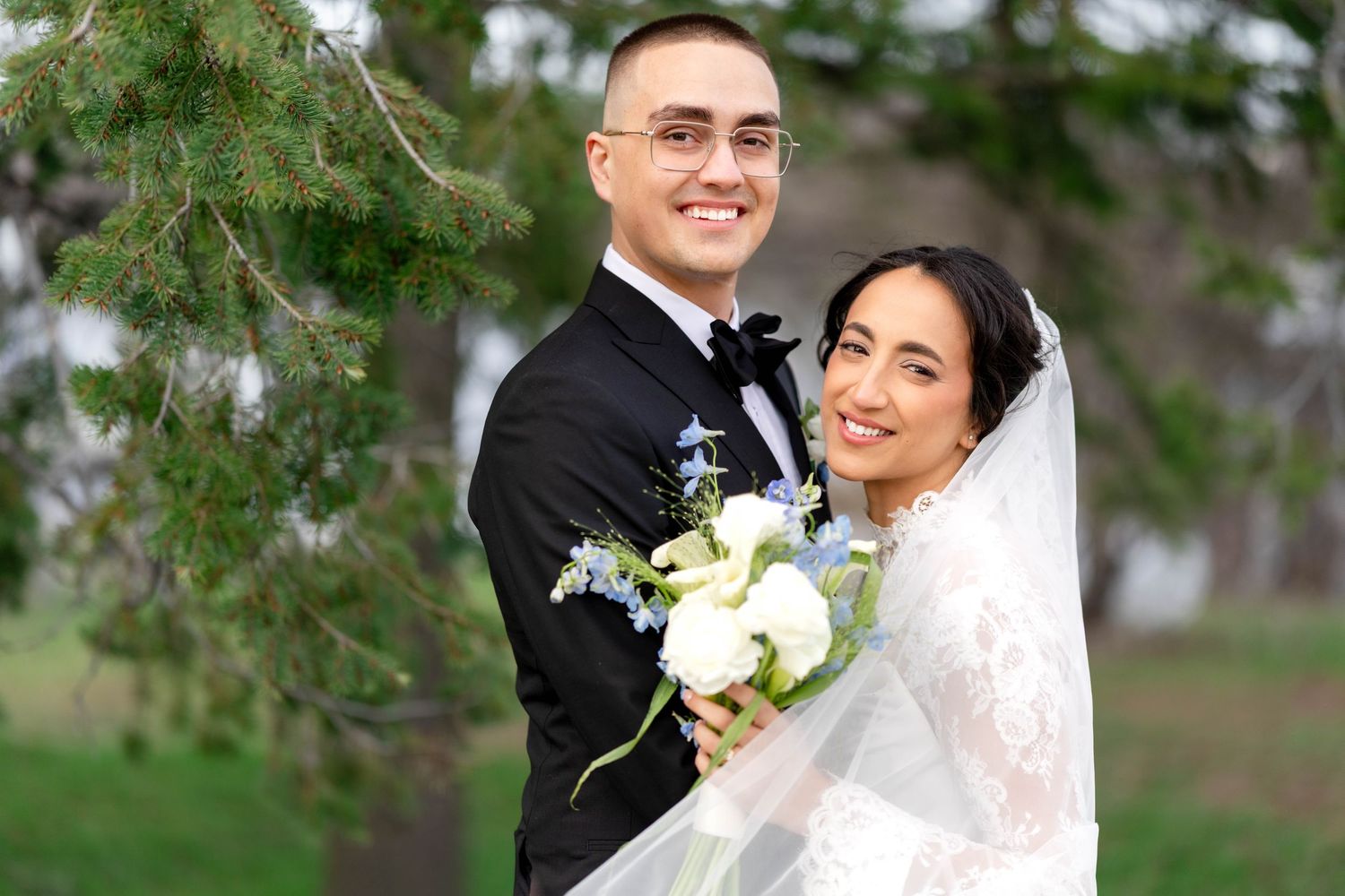 Smiling bride and groom posing outdoors near pine trees, bride holding white floral bouquet with veil flowing.