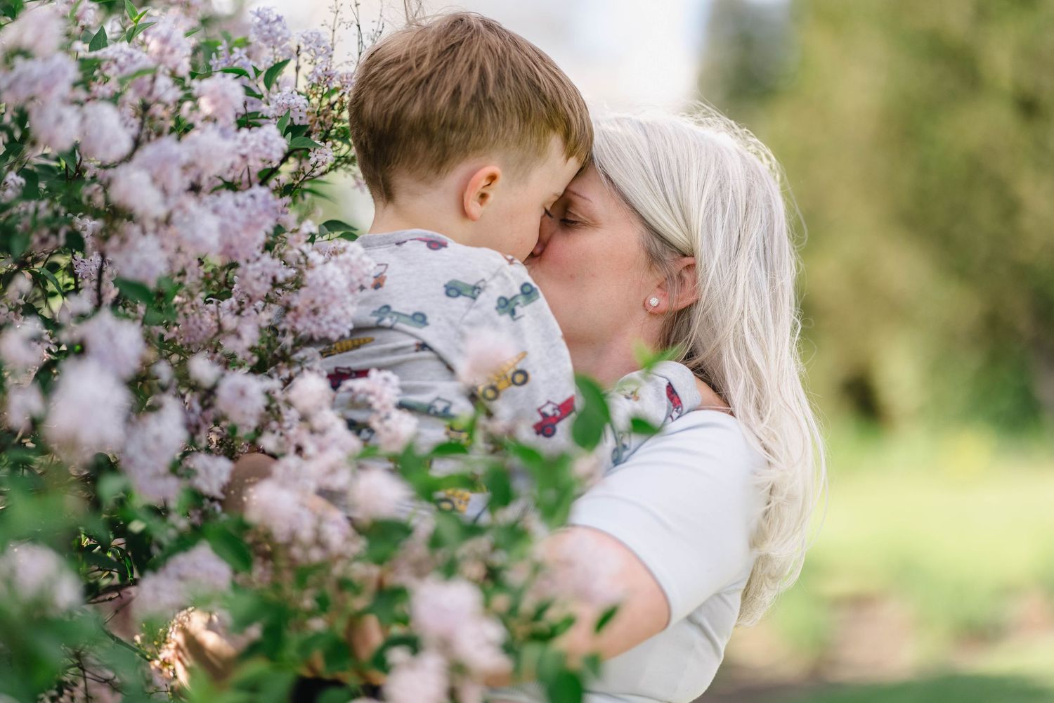 A mother and young son share a tender kiss surrounded by blooming pink flowers in a lush garden.