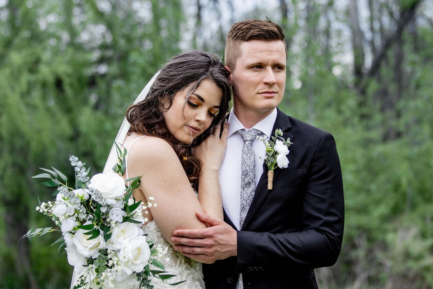 A romantic wedding couple embraces in a lush green forest setting with white floral bouquet details.