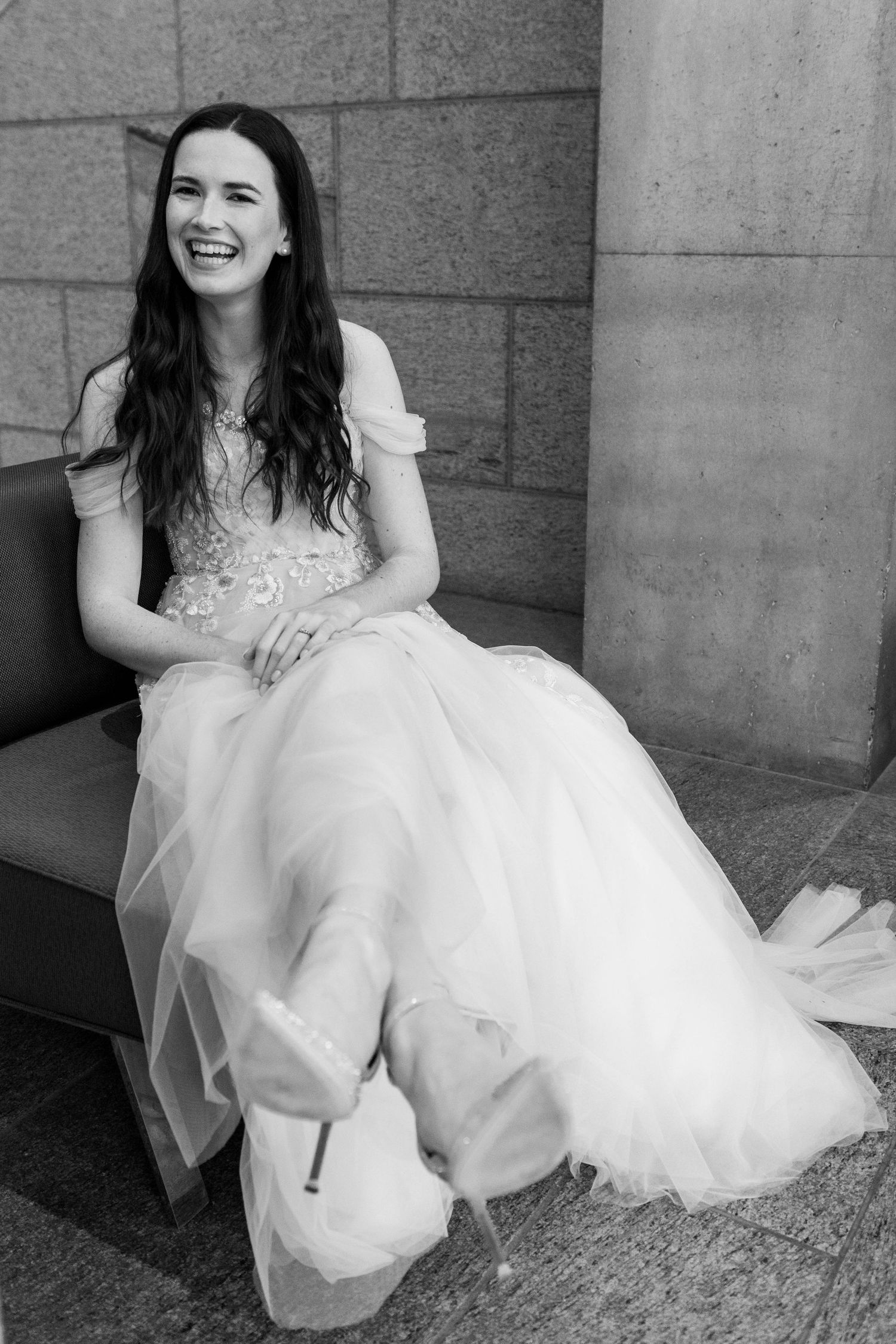 Black and white photo series showing a bride in a flowing white dress sitting and laughing against a stone wall.