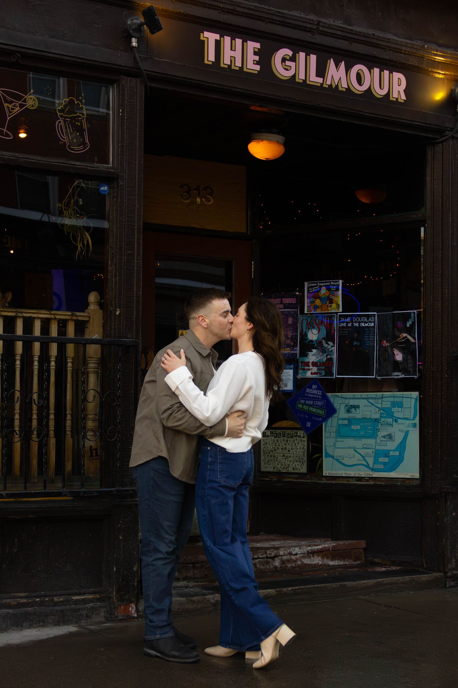 A couple shares a romantic moment outside The Gilmour establishment, with neon signs and window displays visible in the background.