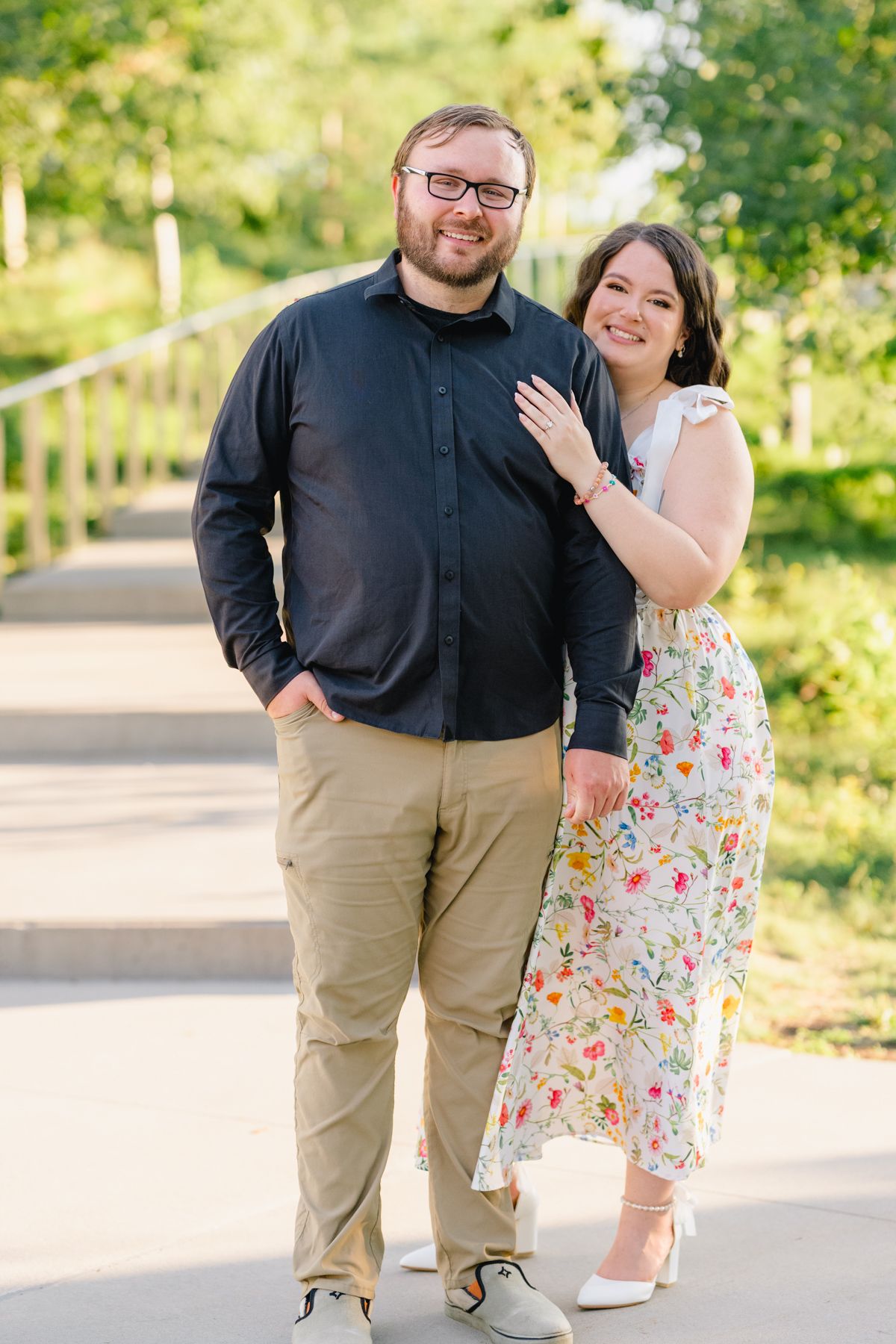 Couple smiling outdoors, man in black shirt and khakis, woman in floral dress, posing on a sunny tree-lined path.