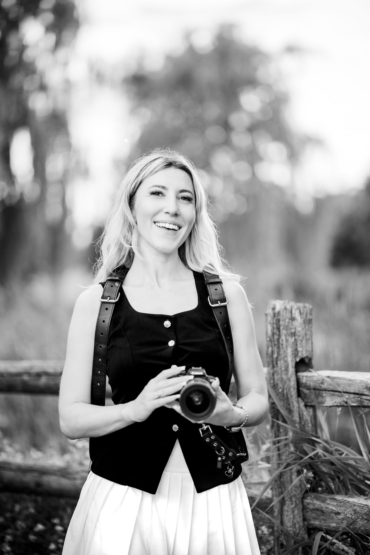 Smiling blonde woman holding a camera outdoors near a wooden fence in a black and white portrait photo.