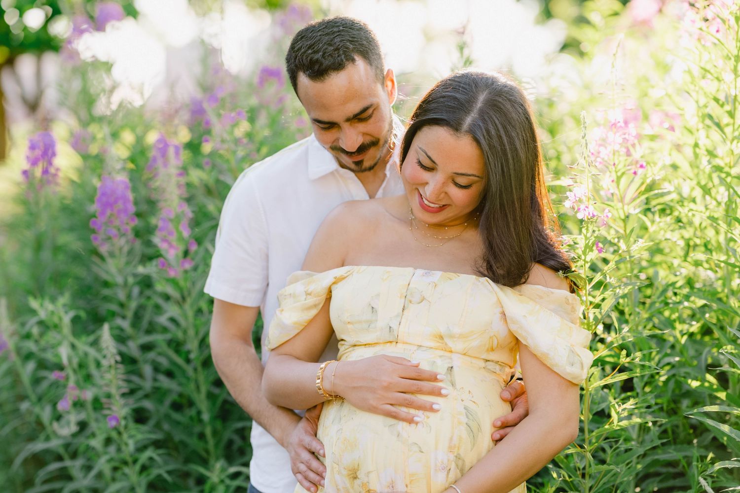 Couple embracing outdoors in a garden, pregnant woman in yellow dress holding baby bump among purple wildflowers.