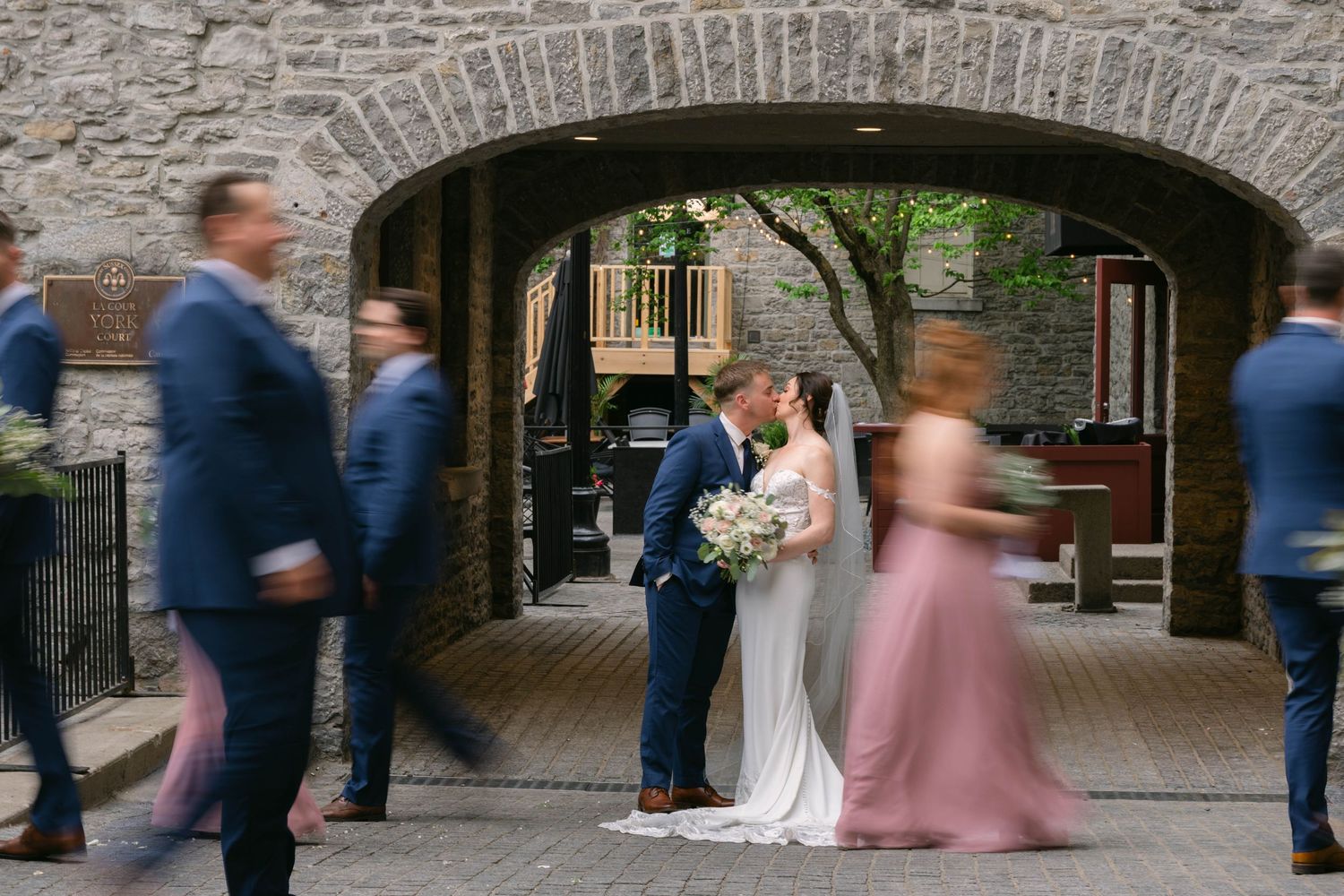 Bride and groom share a kiss under a stone archway while wedding party walks past in blue suits and pink dresses.