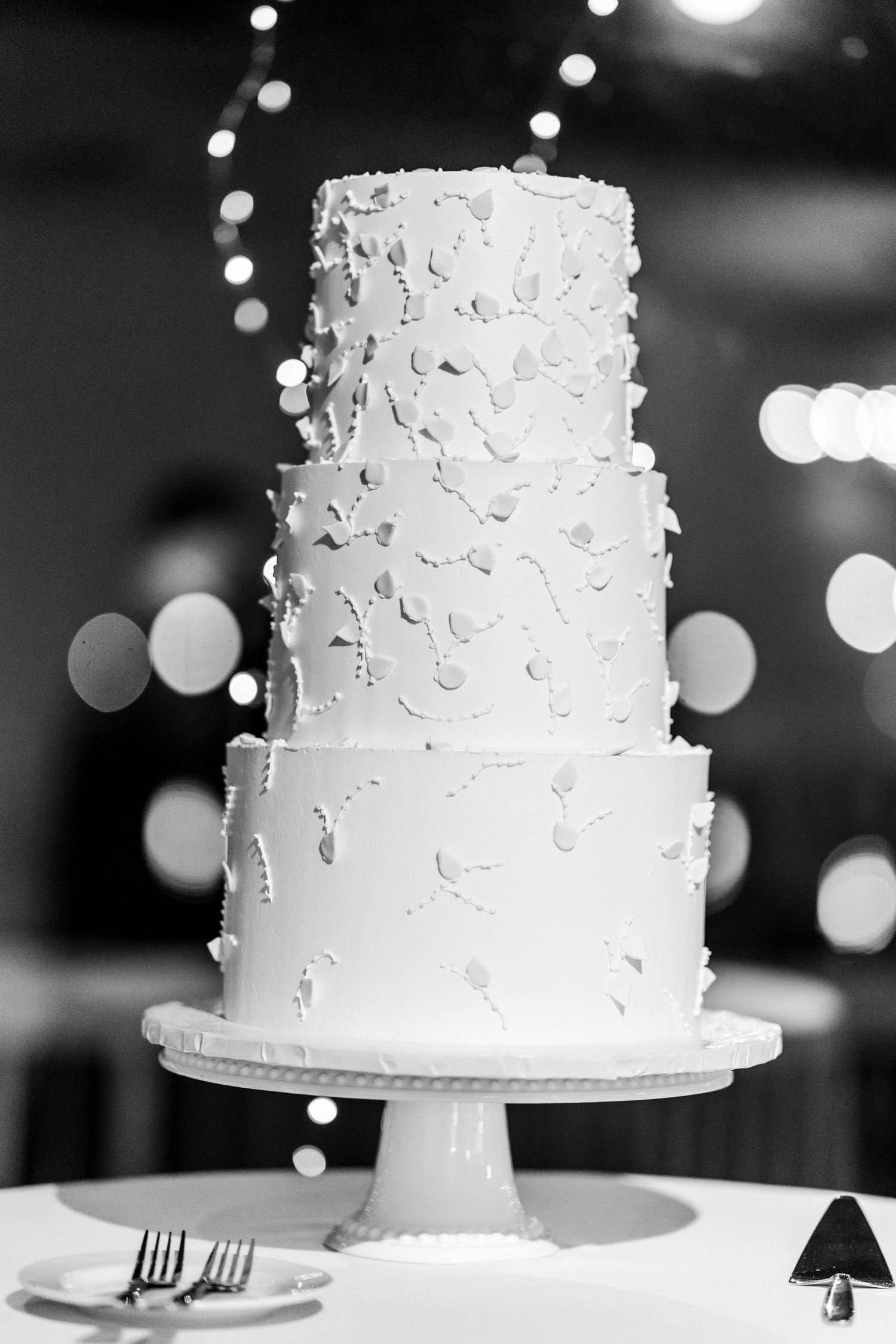 Elegant three-tier white wedding cake with petal decorations displayed on cake stand with bokeh lights.