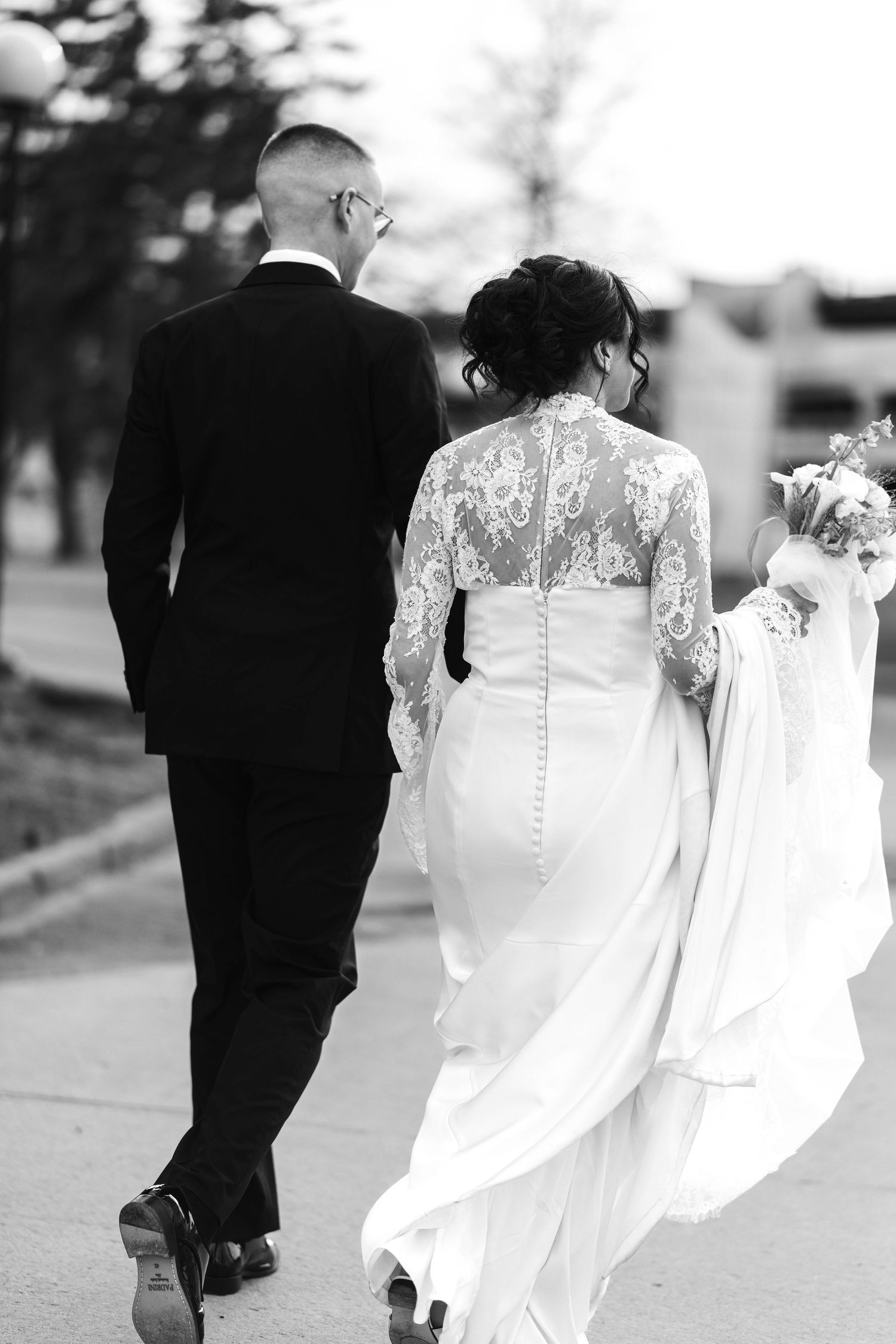 Black and white photo of a bride and groom walking away, bride in lace wedding gown holding bouquet.
