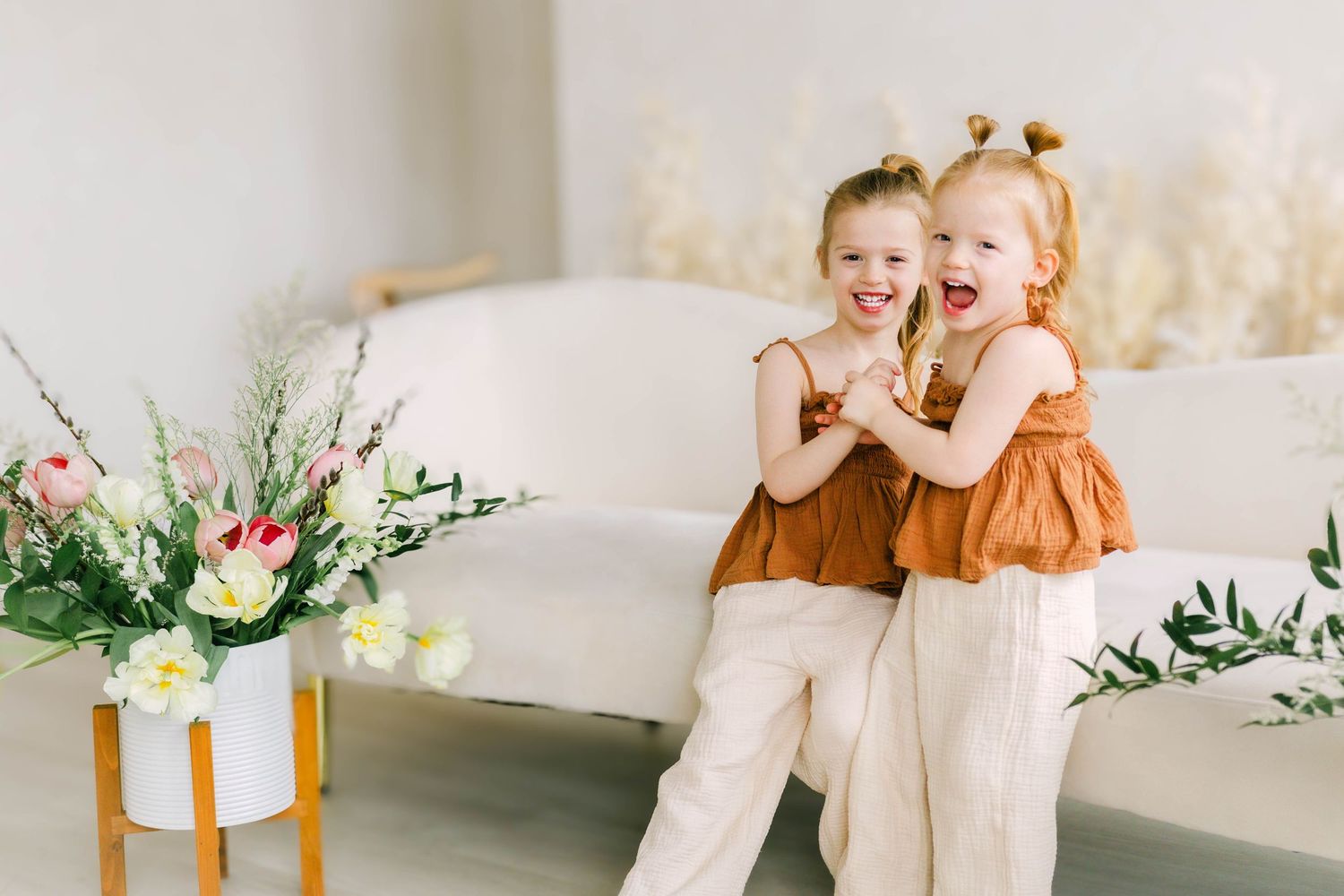Two young children in earth-toned outfits share a playful moment beside floral arrangements in a bright studio setting.