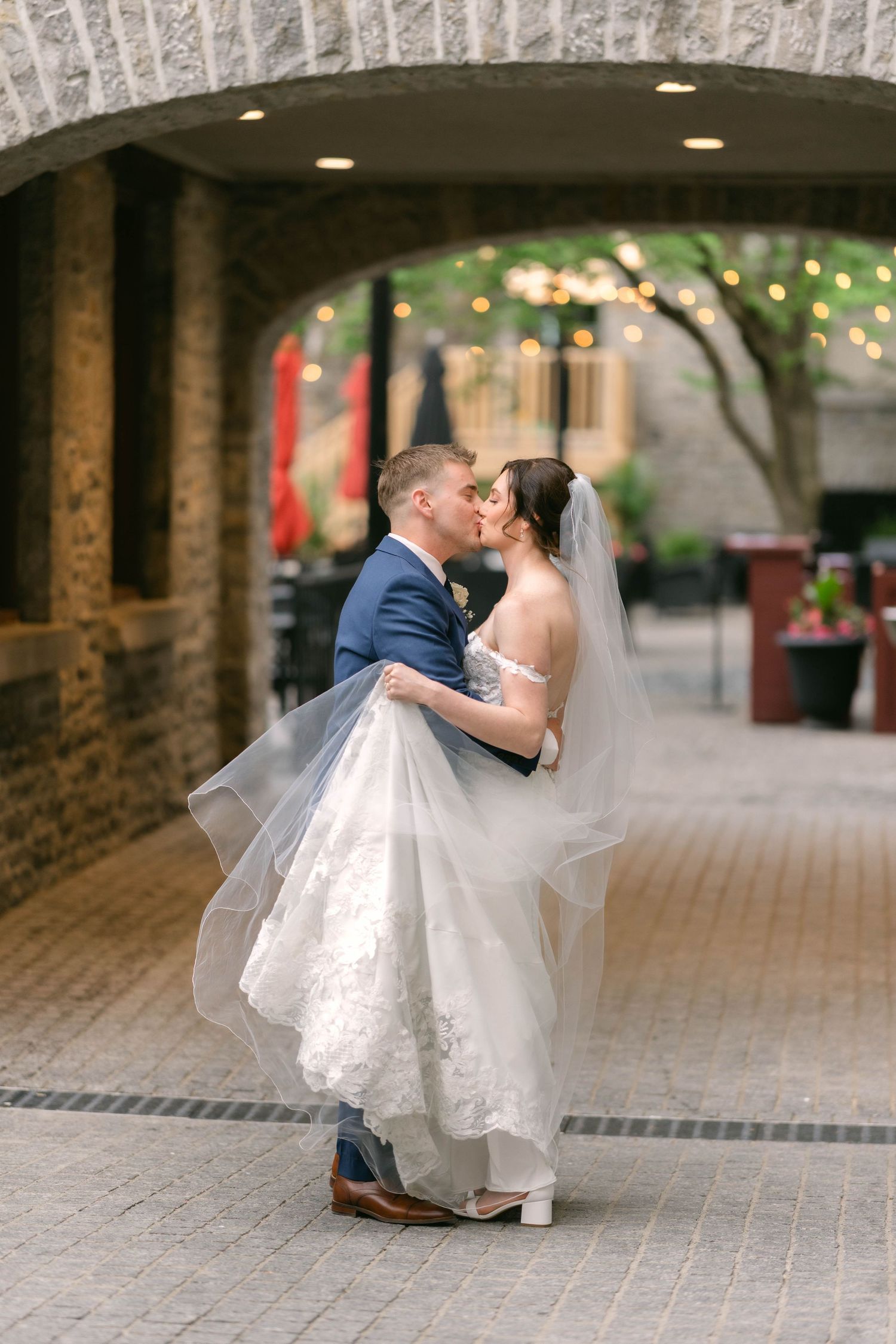 Groom sweeps bride off her feet for a romantic kiss under a stone arch with warm string lights.