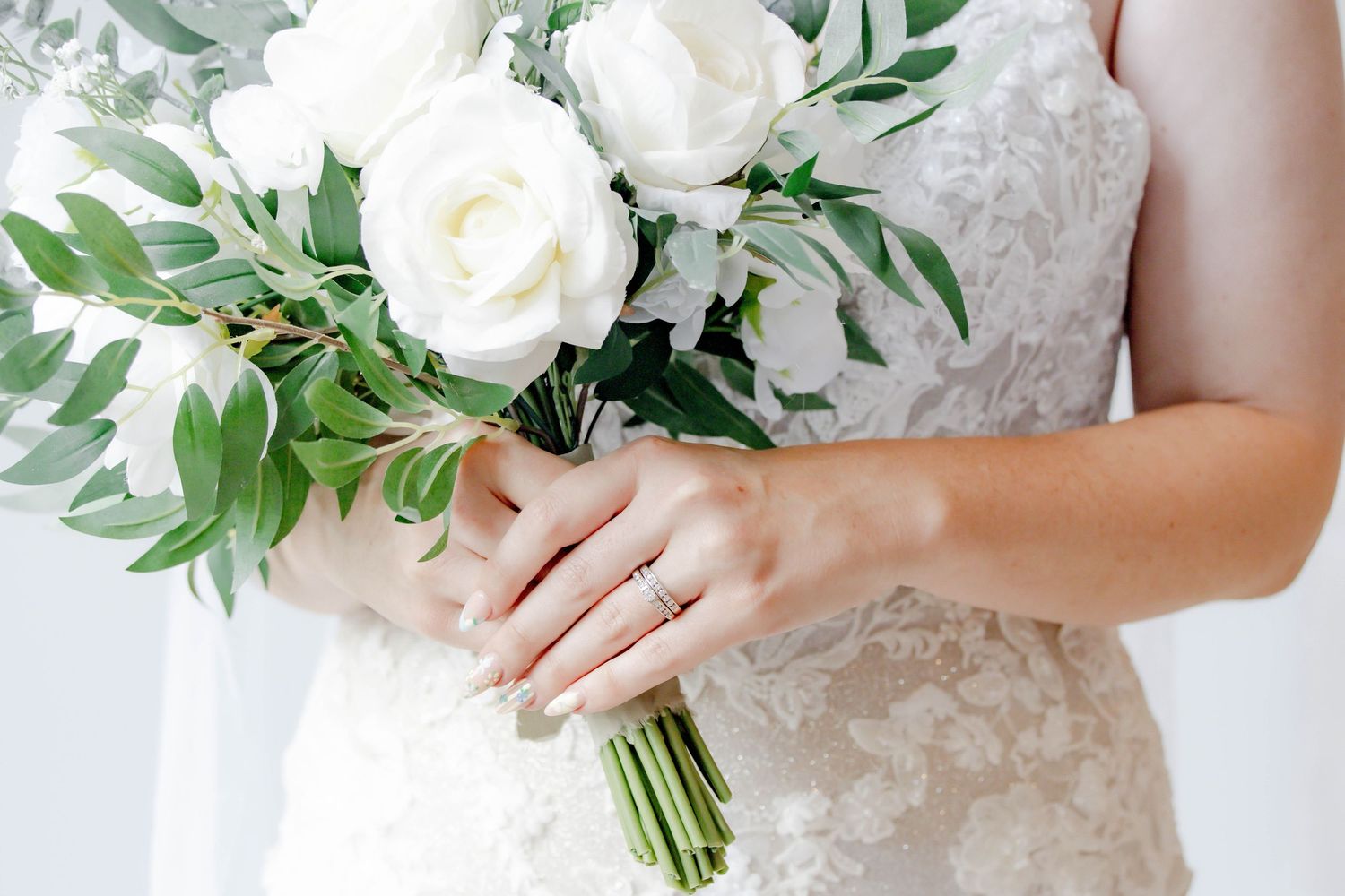 Elegant white bridal bouquet with lush greenery and roses held against a lace wedding dress.