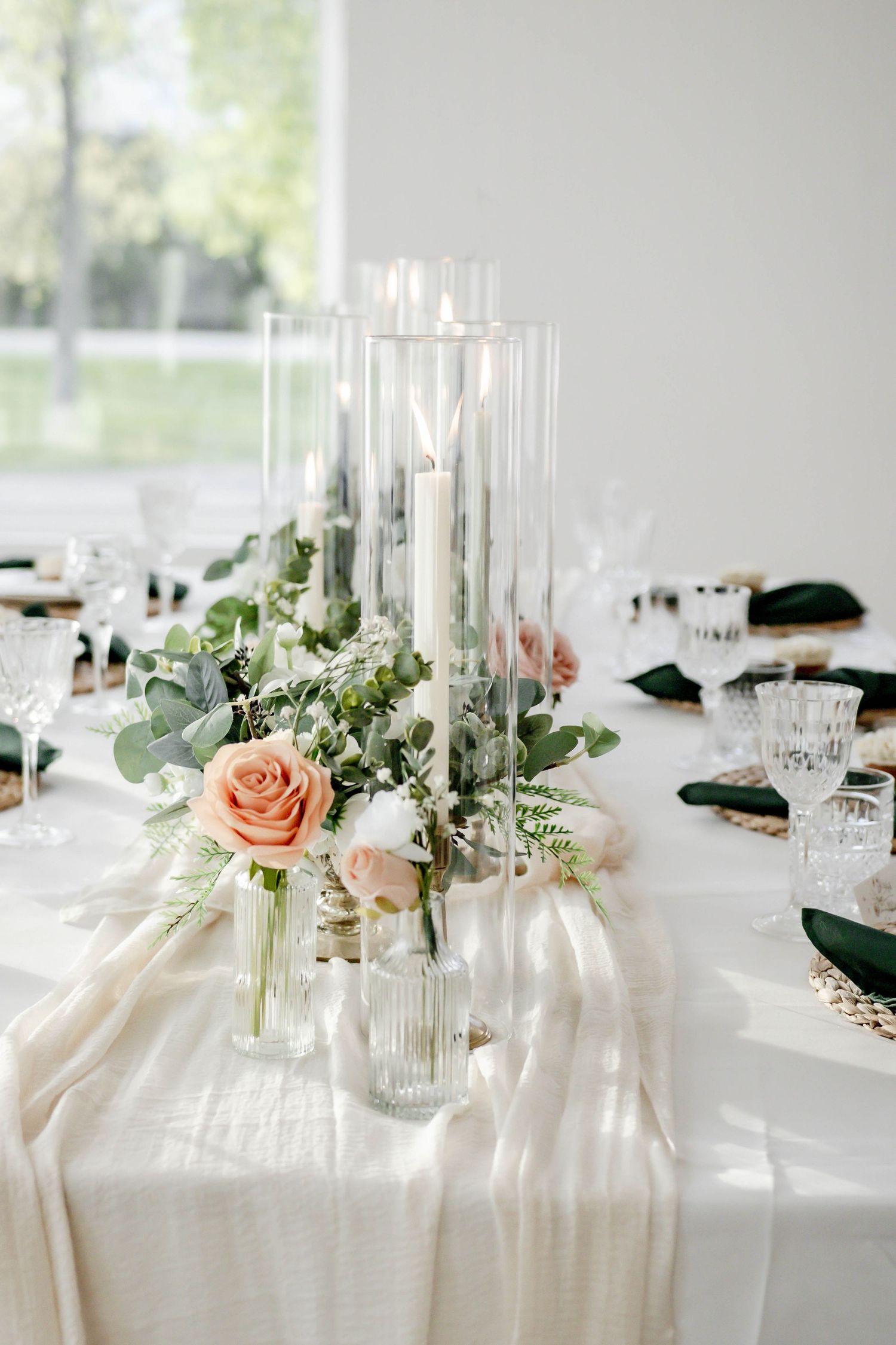 Elegant wedding reception table with blush roses, eucalyptus, and tall glass candle holders on white linens.