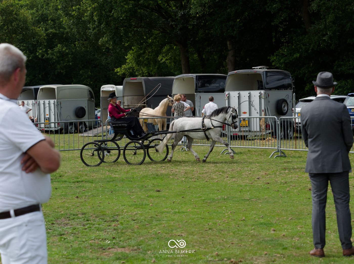 Wide view of a horse show arena with spectators, fencing, and trailers in the background.