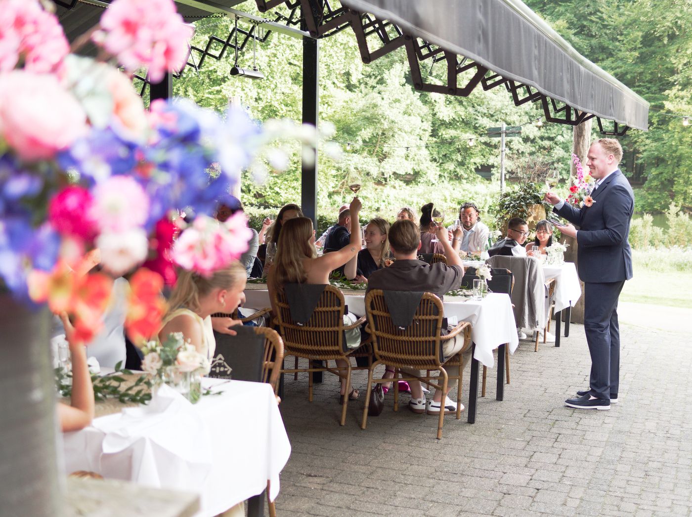Man giving speech at outdoor wedding reception with floral arrangements and guests seated at decorated tables.