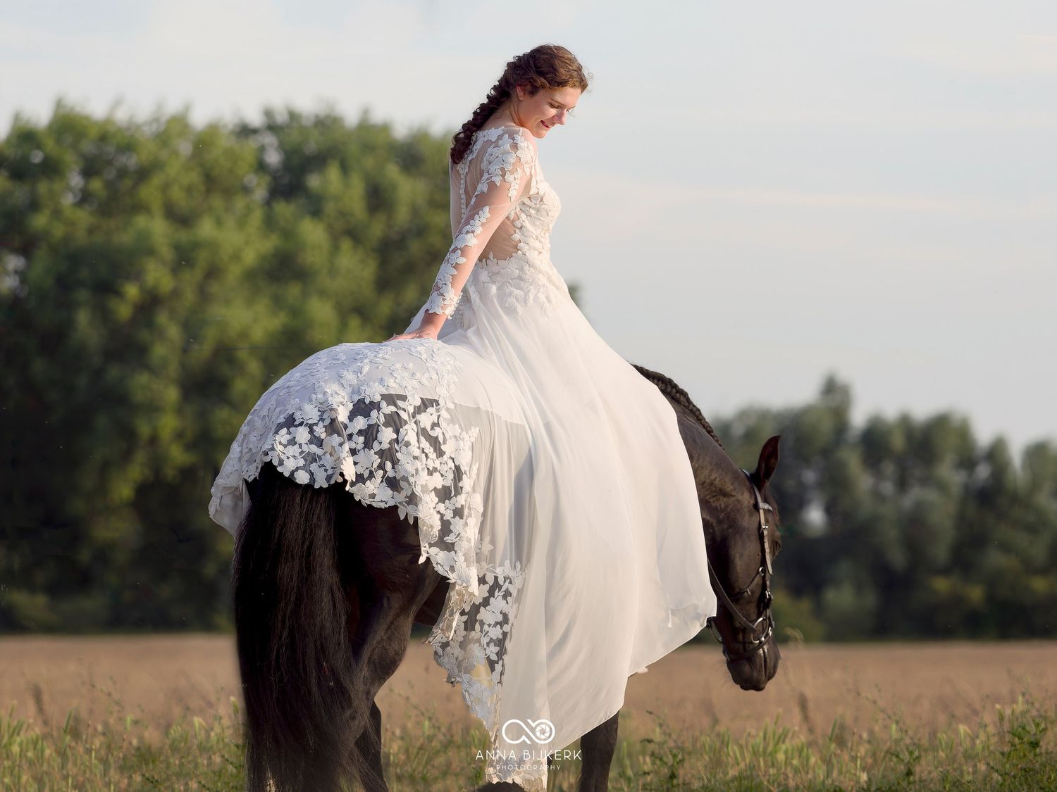 Bride in white lace gown riding a dark horse through a field, with her dress draped elegantly over the horse.