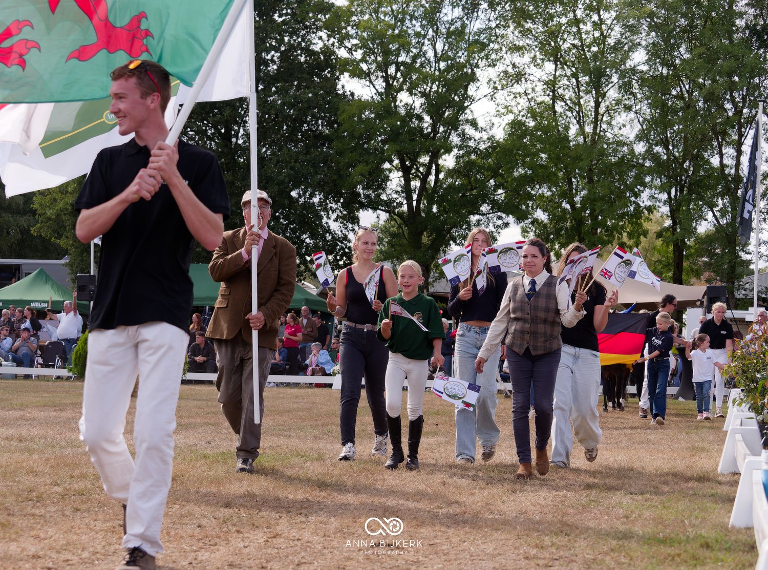 Group of people marching in an outdoor ceremony, led by a young man carrying a Welsh flag.