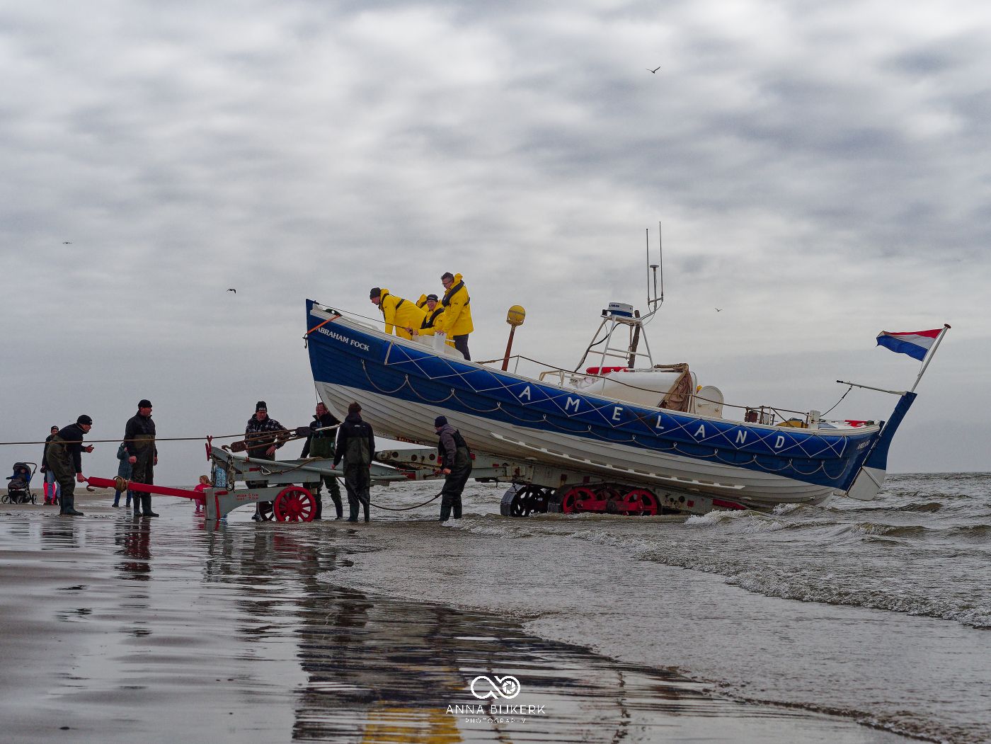 A fishing boat rests on the beach during low tide as people gather around it under cloudy skies.