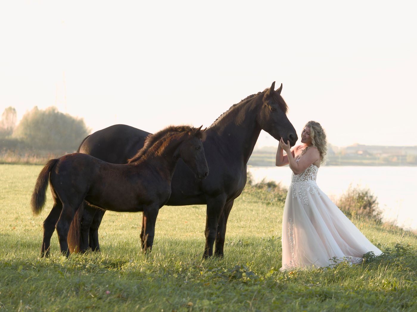 Woman in white gown stands with two dark horses in a sunlit green field near a tranquil lake.