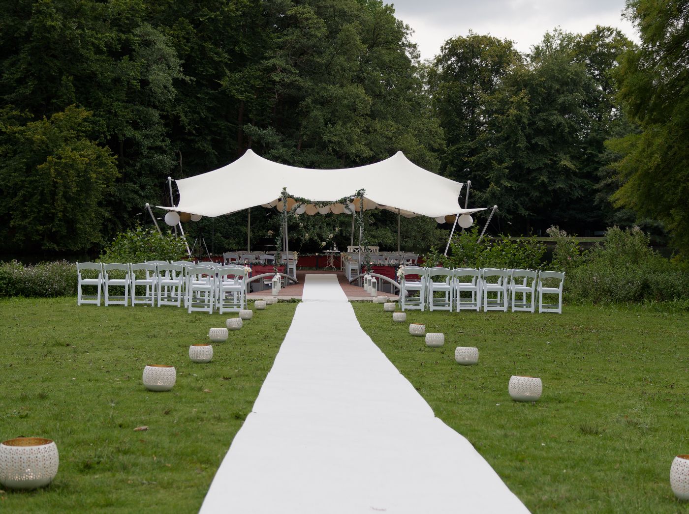 Outdoor wedding ceremony setup with white aisle runner, chairs, and elegant stretch tent canopy surrounded by lush greenery.