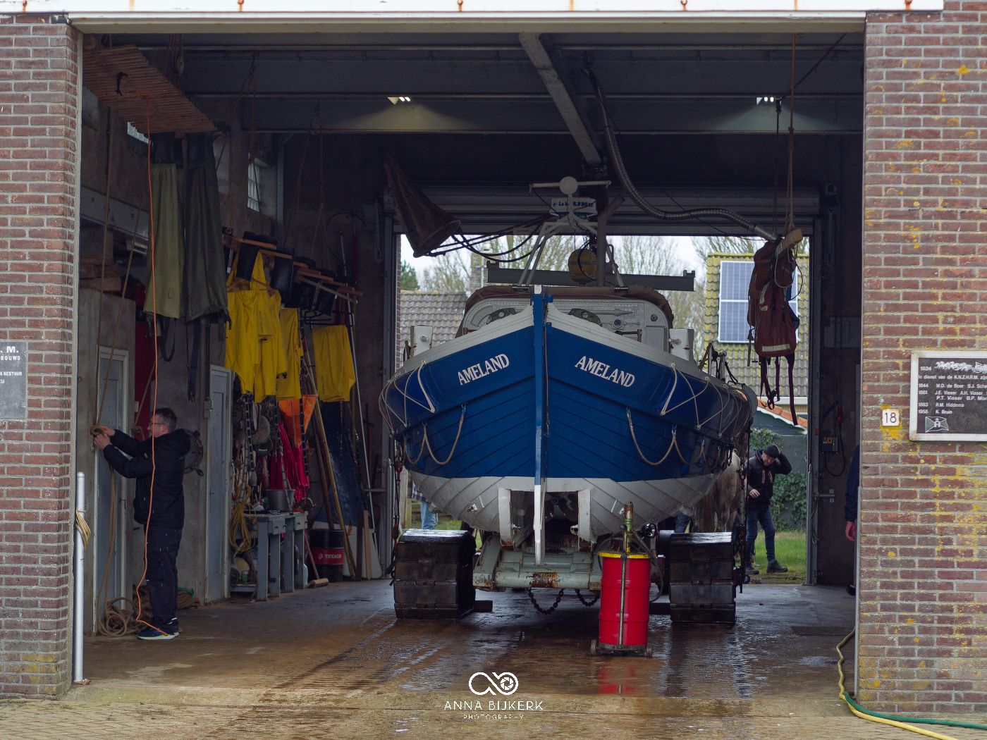 A blue boat on a trailer is parked inside a brick garage with yellow clothing hanging on the wall.
