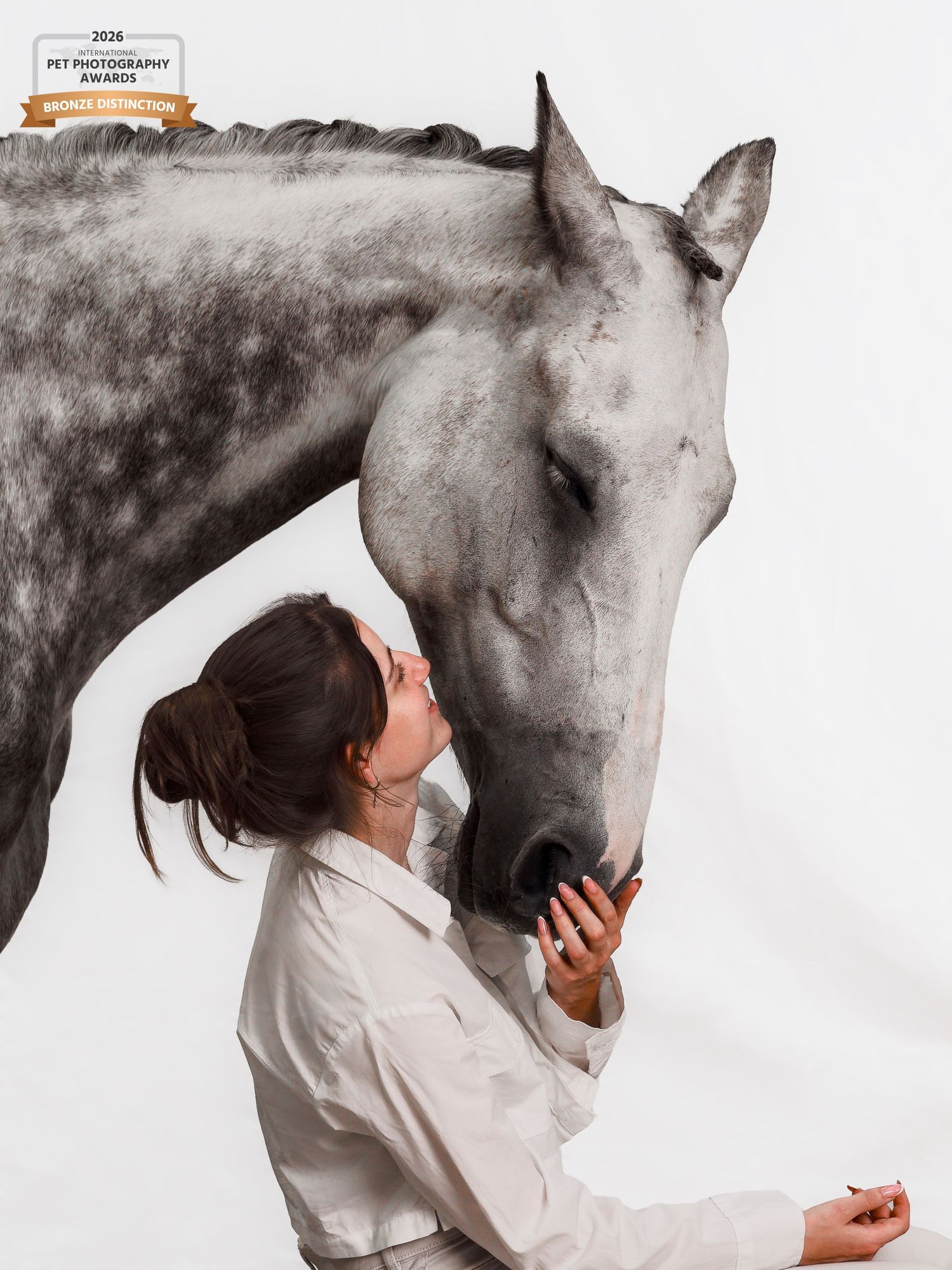 Award-winning photo of a woman in white gently bonding with a dapple gray horse against a white background.