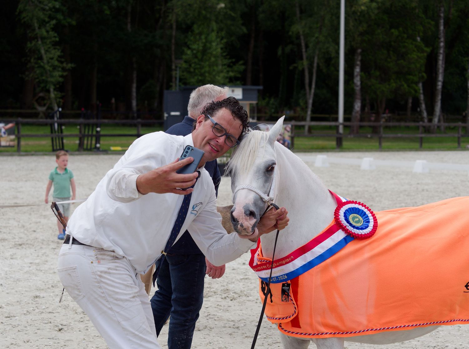 Man taking selfie with award-winning white horse wearing blue and red rosette at equestrian competition.