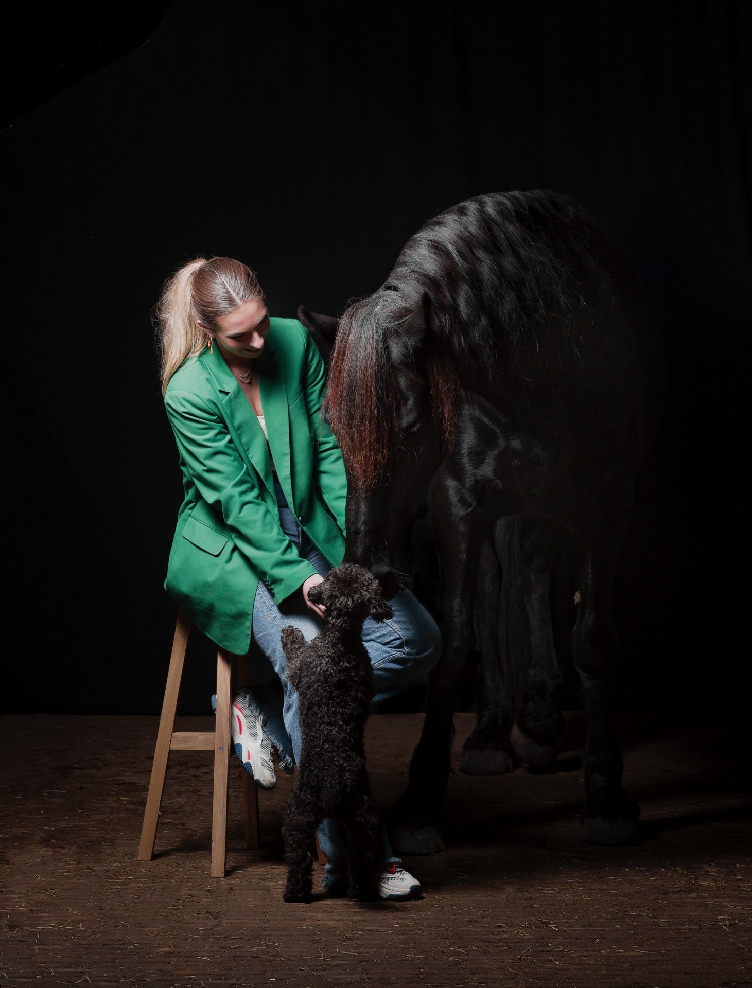 A person in a green jacket sits hunched over on a wooden stool against a dark background in a dramatic pose.