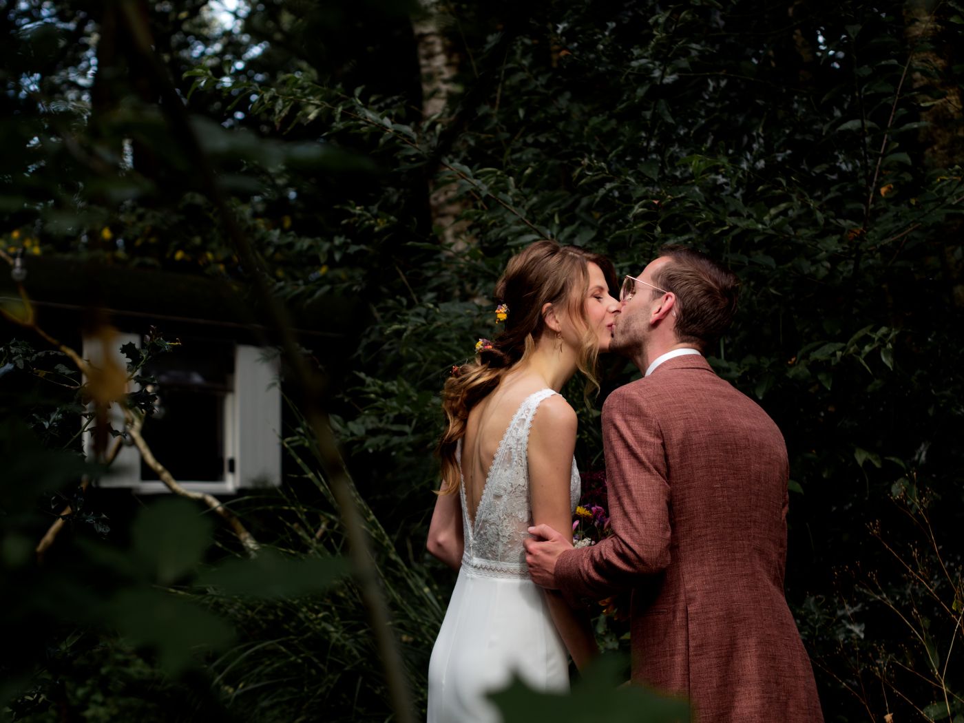A bride and groom share a romantic kiss surrounded by lush green trees in an outdoor woodland wedding setting.